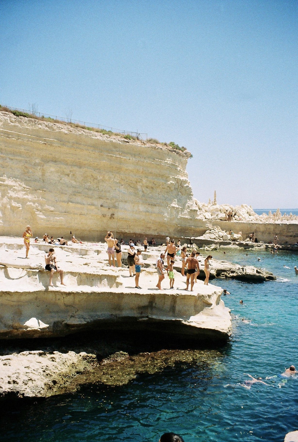 People sunbathing and swimming on white rocky cliffs at a beach by clear blue water, with a large cliff and bright sky in the background.