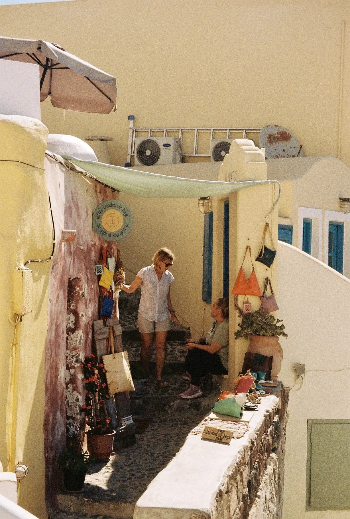 Two women in a small shop on a narrow street, with colorful bags and accessories hanging on the wall, and various items displayed on a stone ledge.