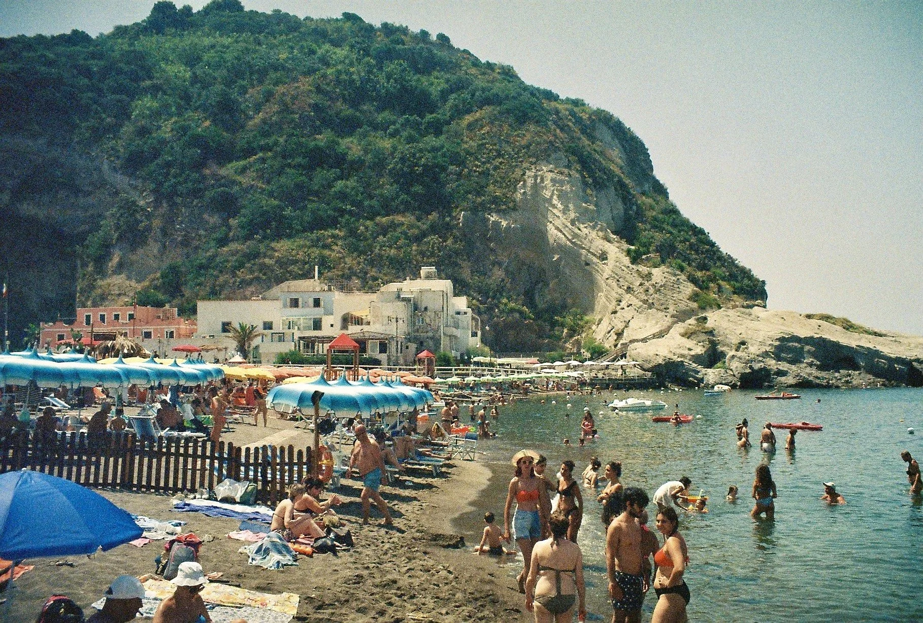 Crowded beach with umbrellas, people swimming, and a hillside with greenery and rocks in the background.