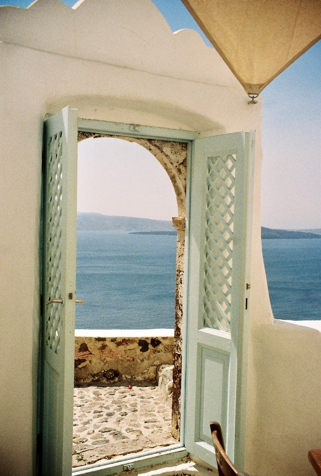View through open pastel green shutters of a white-washed building looking out to a blue sea and distant islands.