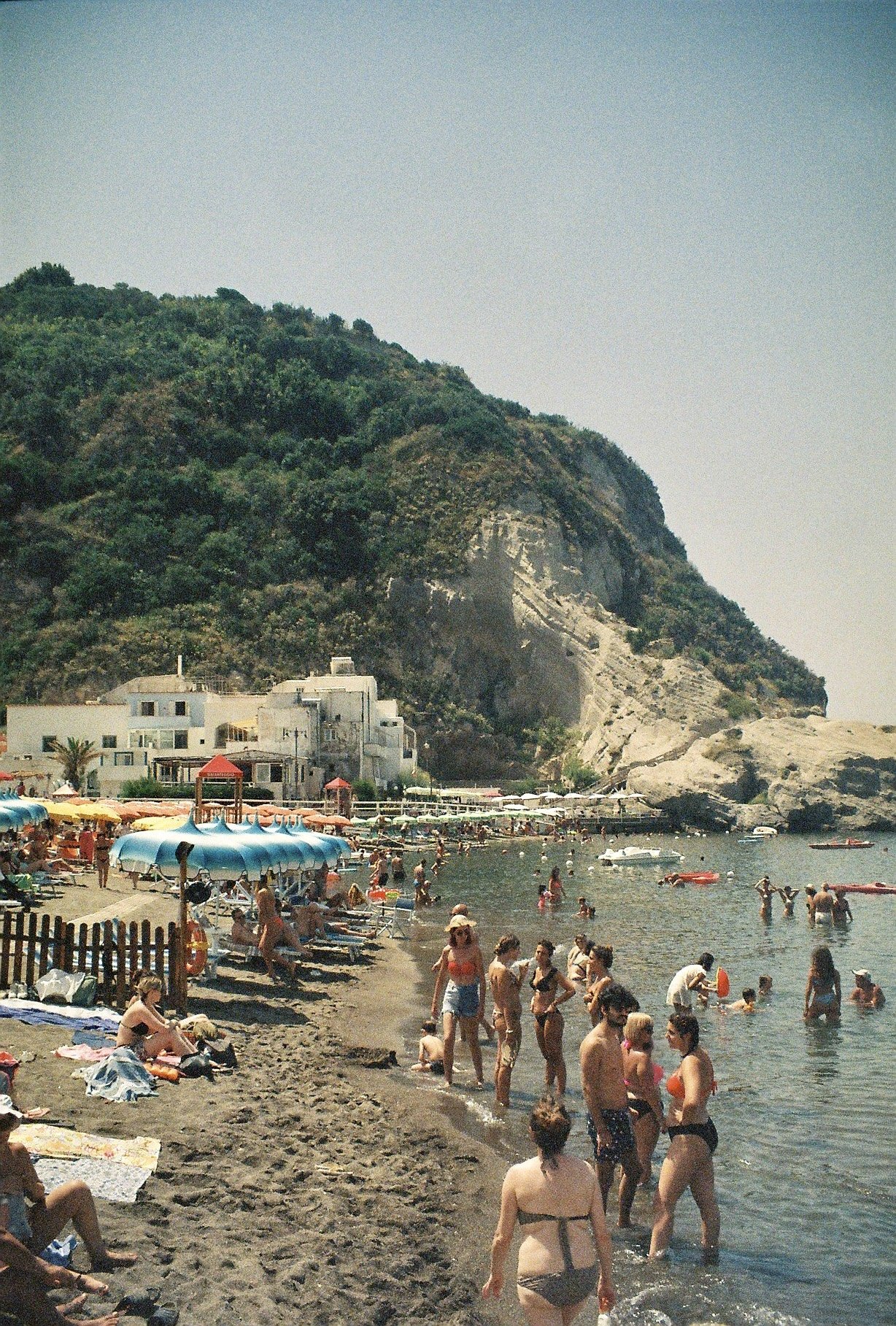 Beach scene with people swimming, sunbathing, and walking along the shore. There are umbrellas and lounge chairs, a rocky hillside covered with greenery, and buildings near the beach. The sky is clear with no clouds.