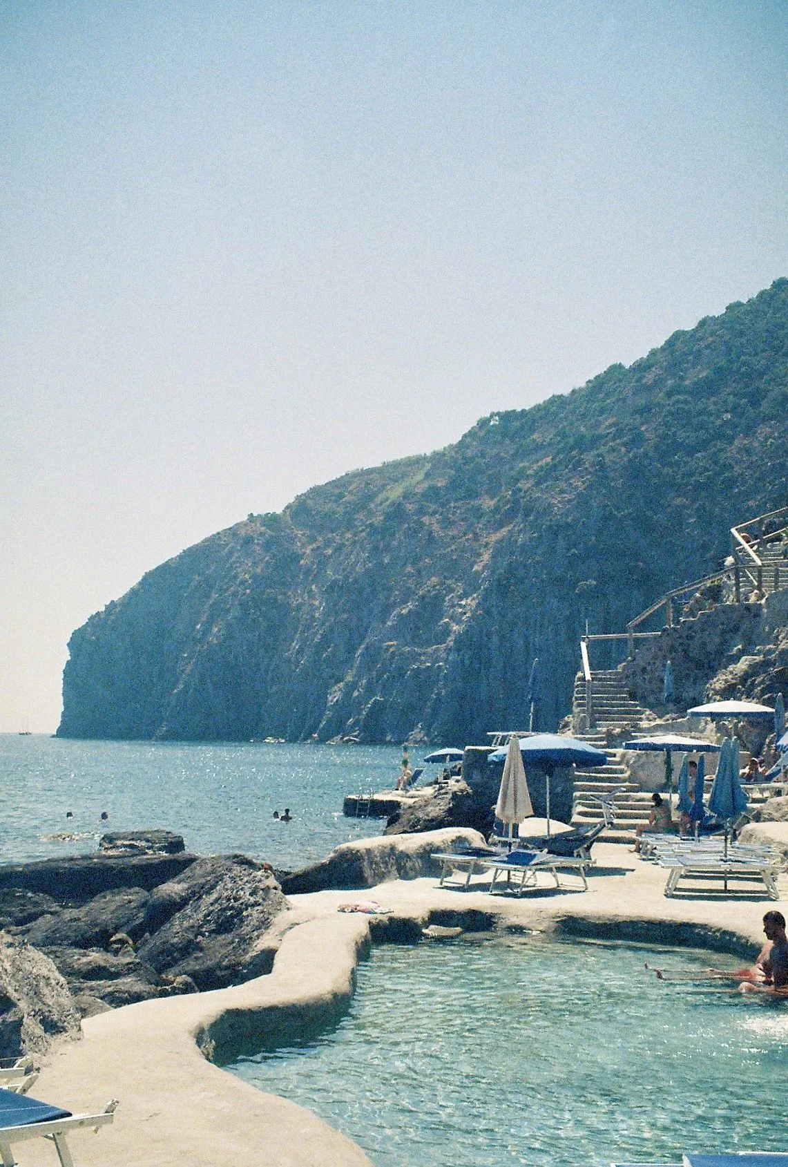 Beach with rocks, umbrellas, and lounge chairs near the ocean, with a large cliff in the background and people swimming.