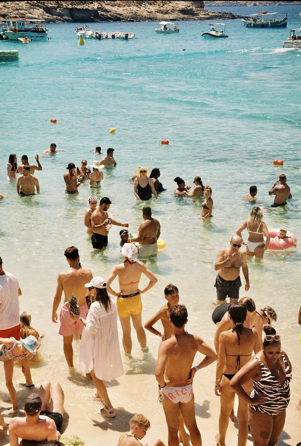 People enjoying a crowded beach with clear blue water, some are swimming while others are standing or sitting on the sandy shore. Many are wearing swimsuits, sun hats, and sunglasses. Small boats are anchored near the shoreline, and a rocky cliff is 
