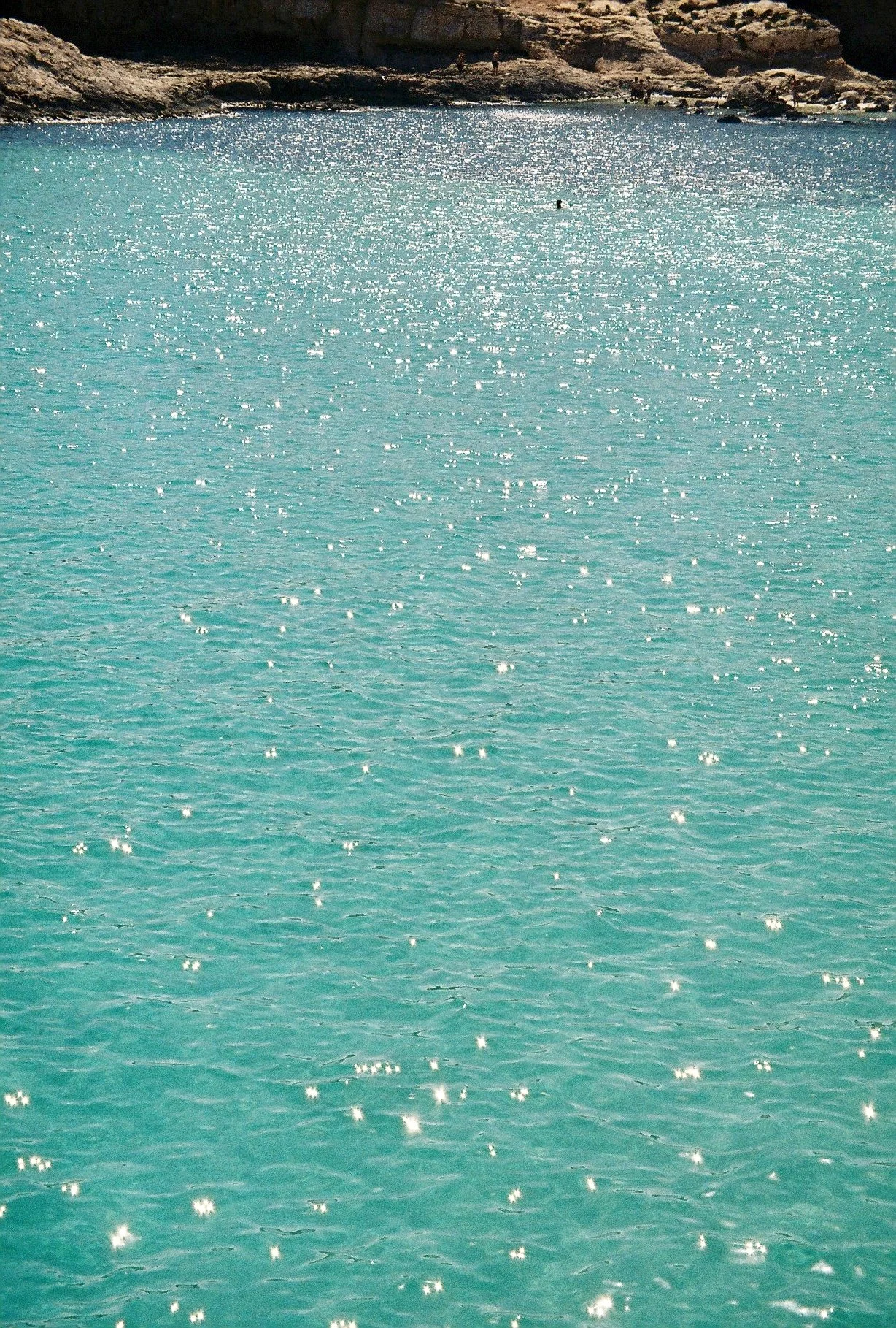 A body of turquoise water reflecting sunlight with rocky cliffs and sparse vegetation in the background.