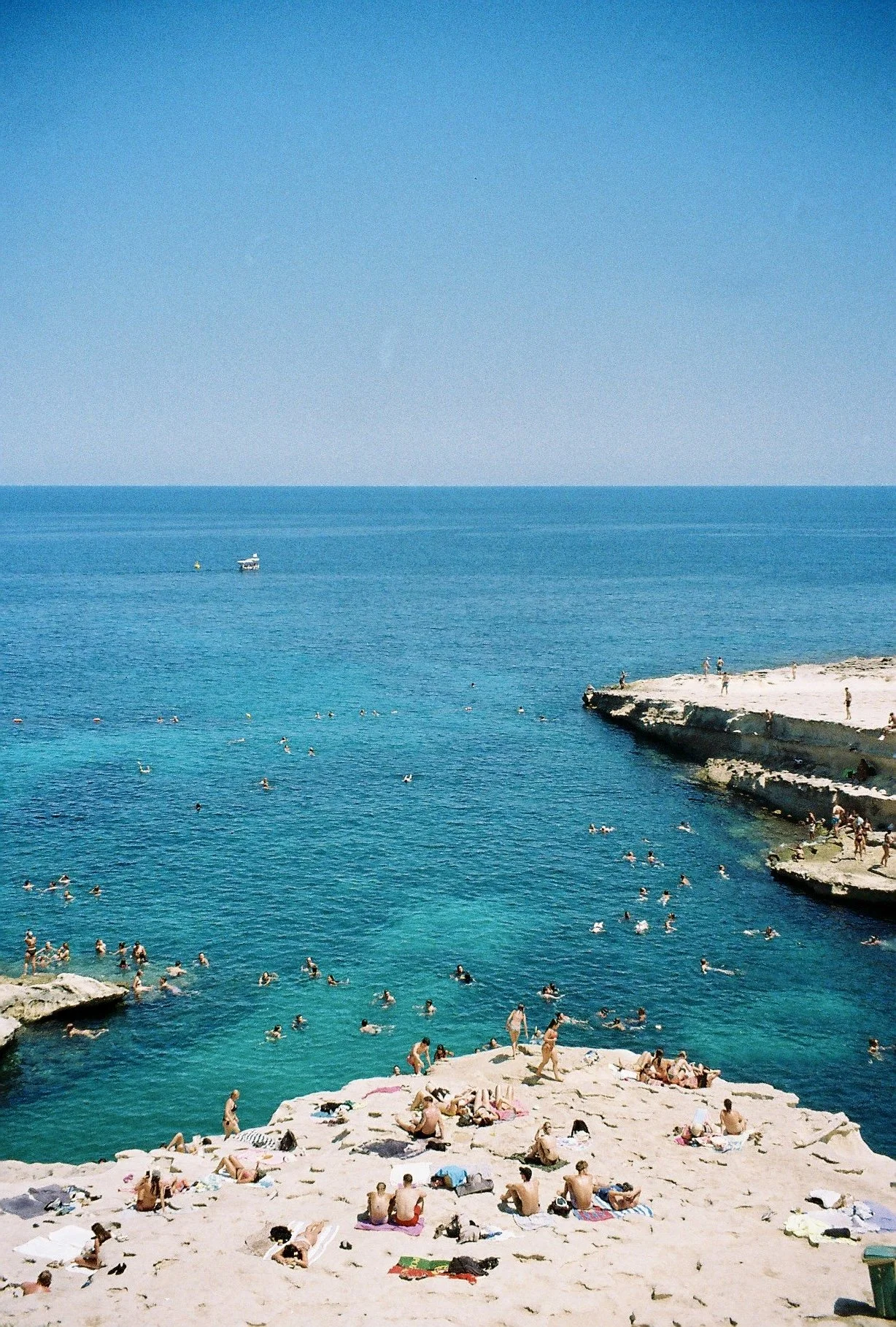 People sunbathing on rocks and swimming in the blue ocean near a rocky coastline under clear sky.