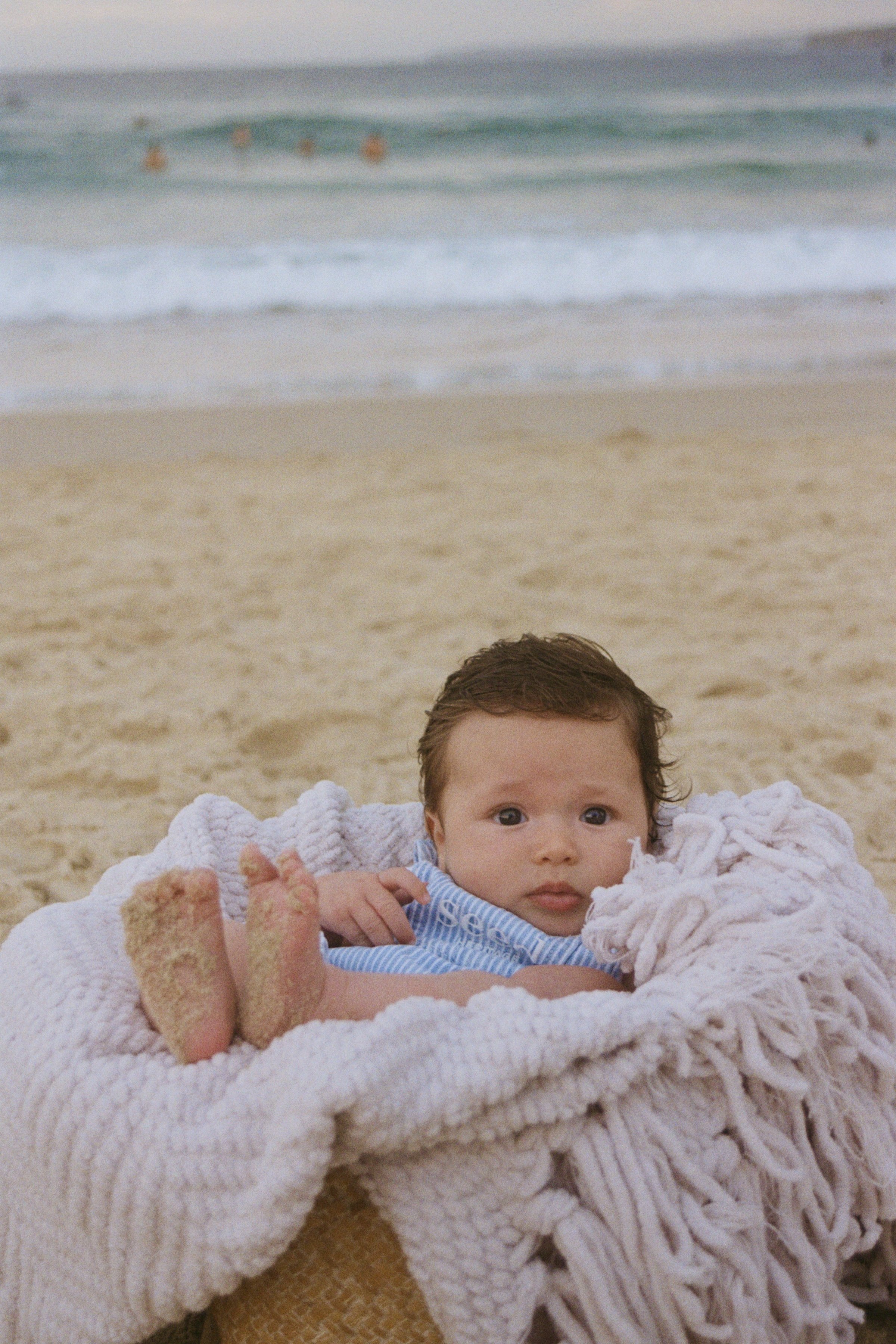 A baby lying on a cream-colored, fluffy blanket at the beach with sand and ocean in the background; part of an adult's foot is visible in the foreground.