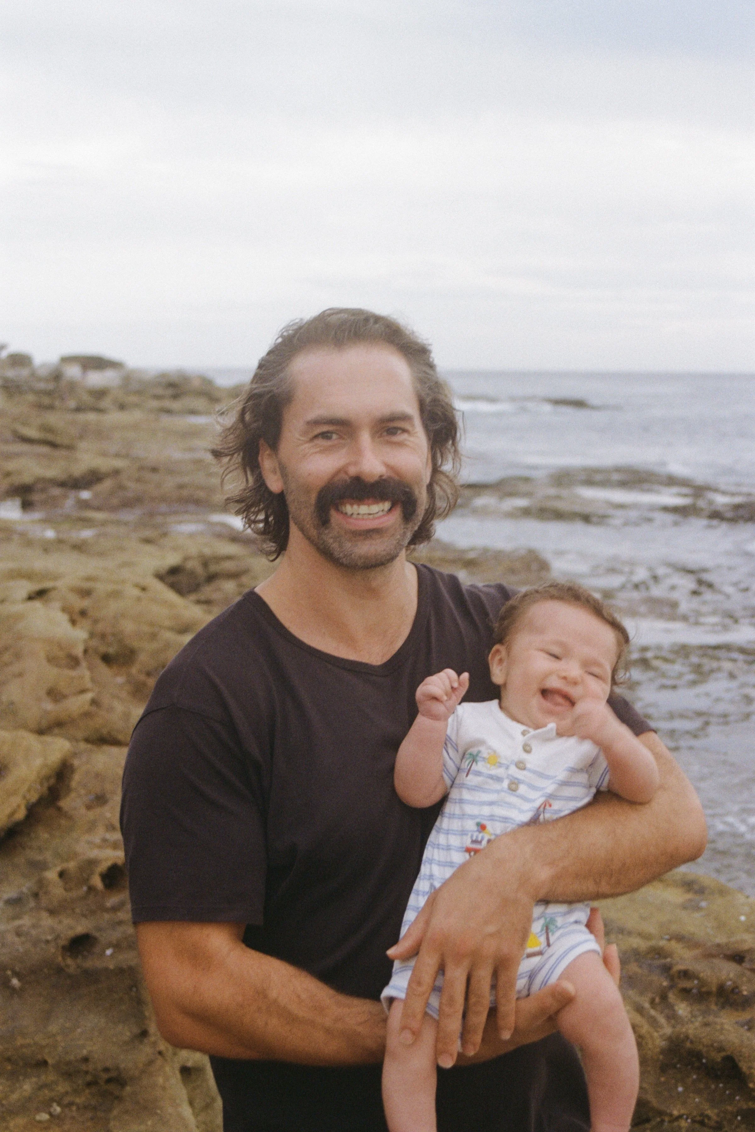 A man with long hair and a mustache holding a smiling baby girl at the beach.