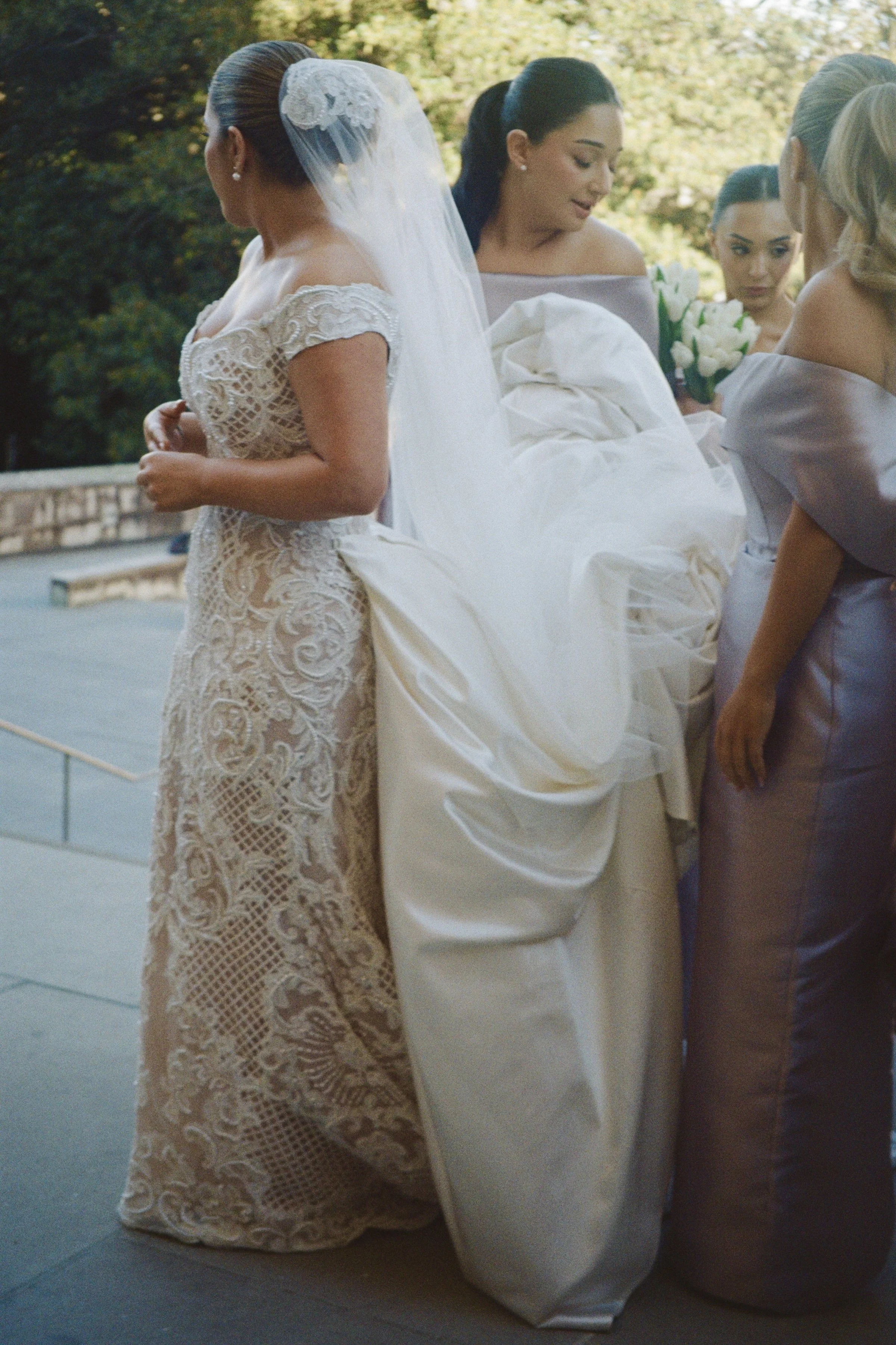 Bridal party standing outdoors, including a bride in a lace wedding dress with a veil and lace details, and bridesmaids holding bouquets of white roses, dressed in lavender satin gowns.