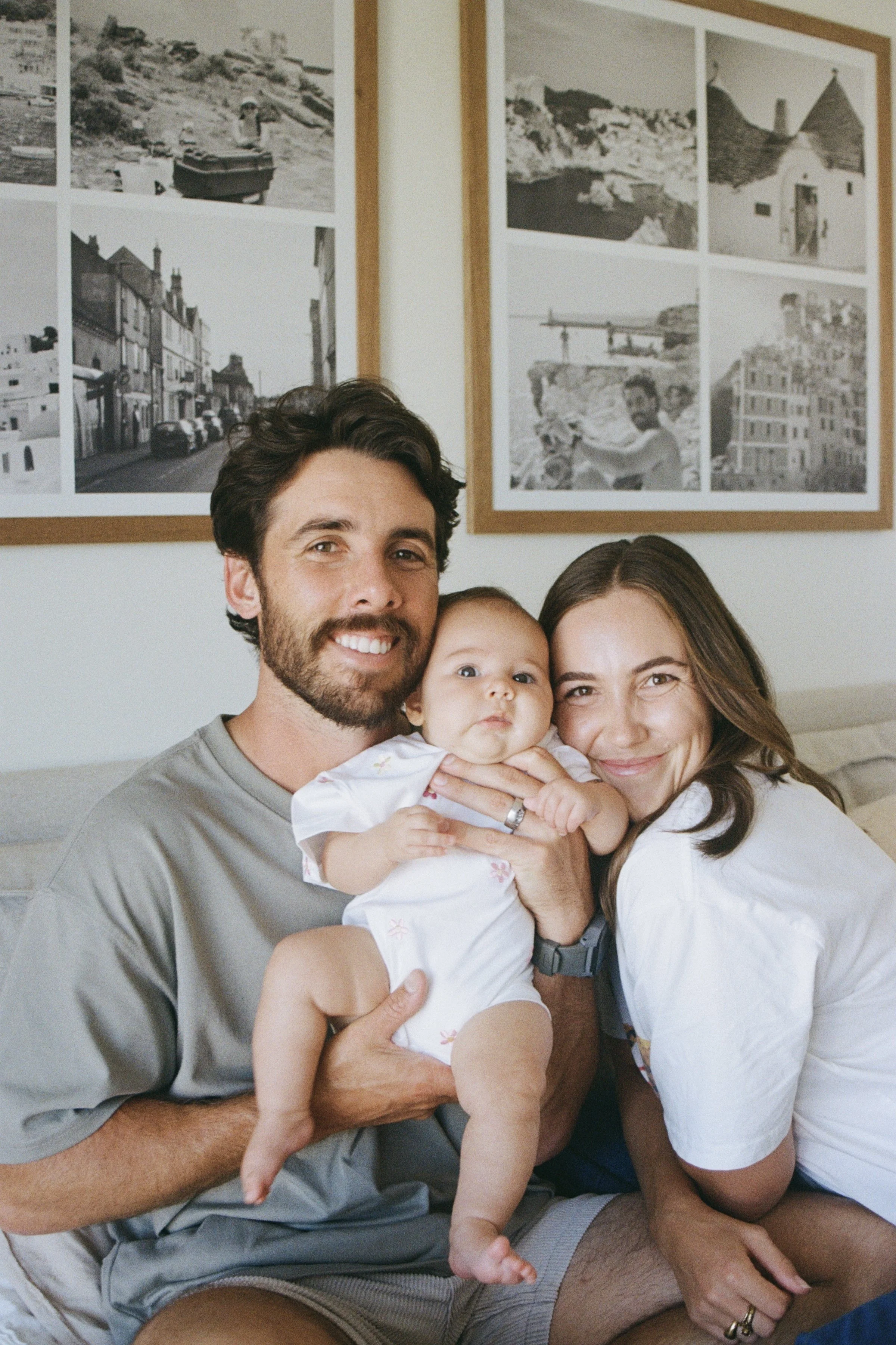A happy family of three, including a man, a woman, and a baby, sitting together indoors with framed black-and-white photographs hanging on the wall behind them.