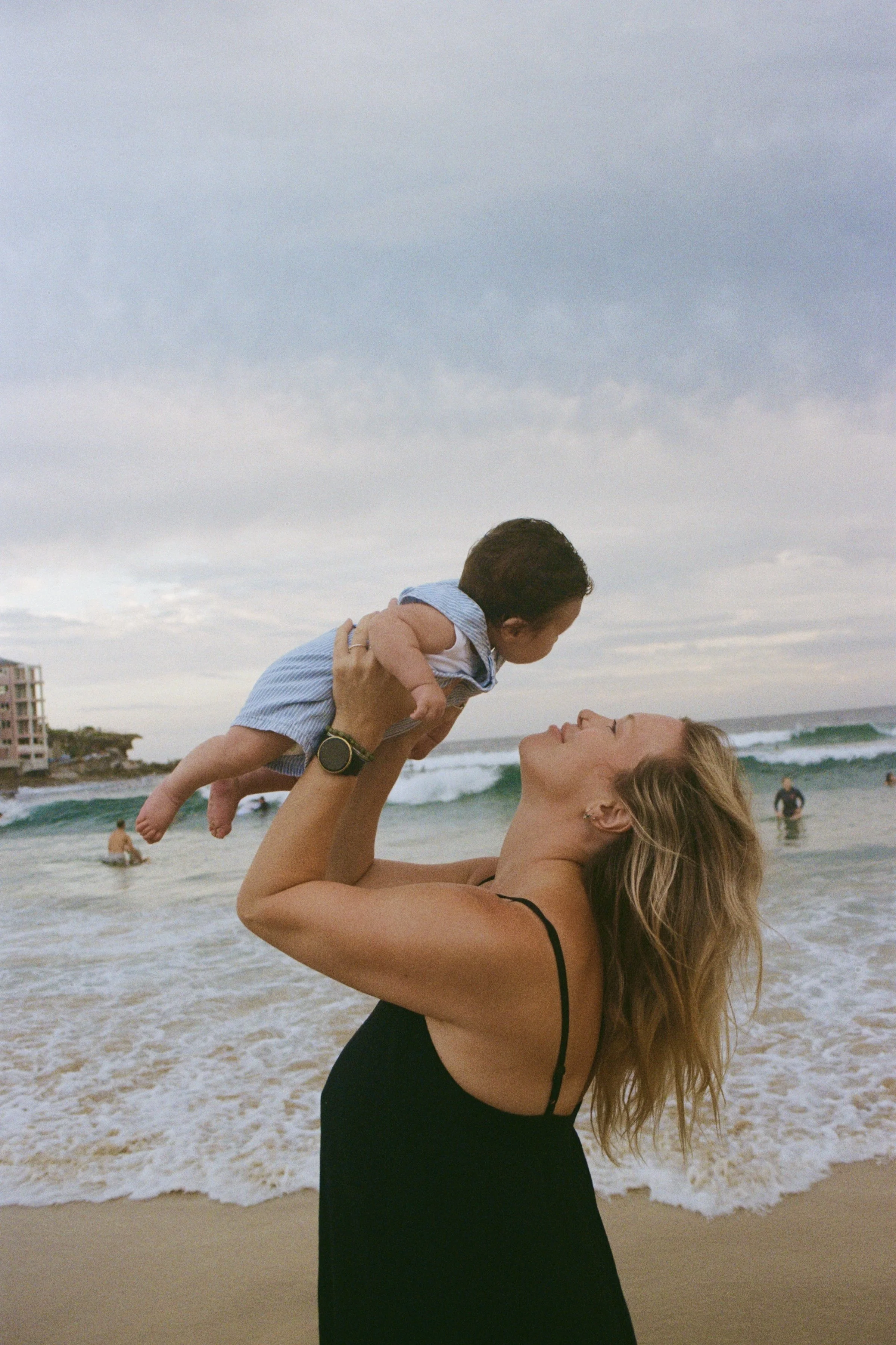 A woman in a black dress lifts a young boy in a striped shirt at the beach, with ocean waves and other beachgoers in the background.
