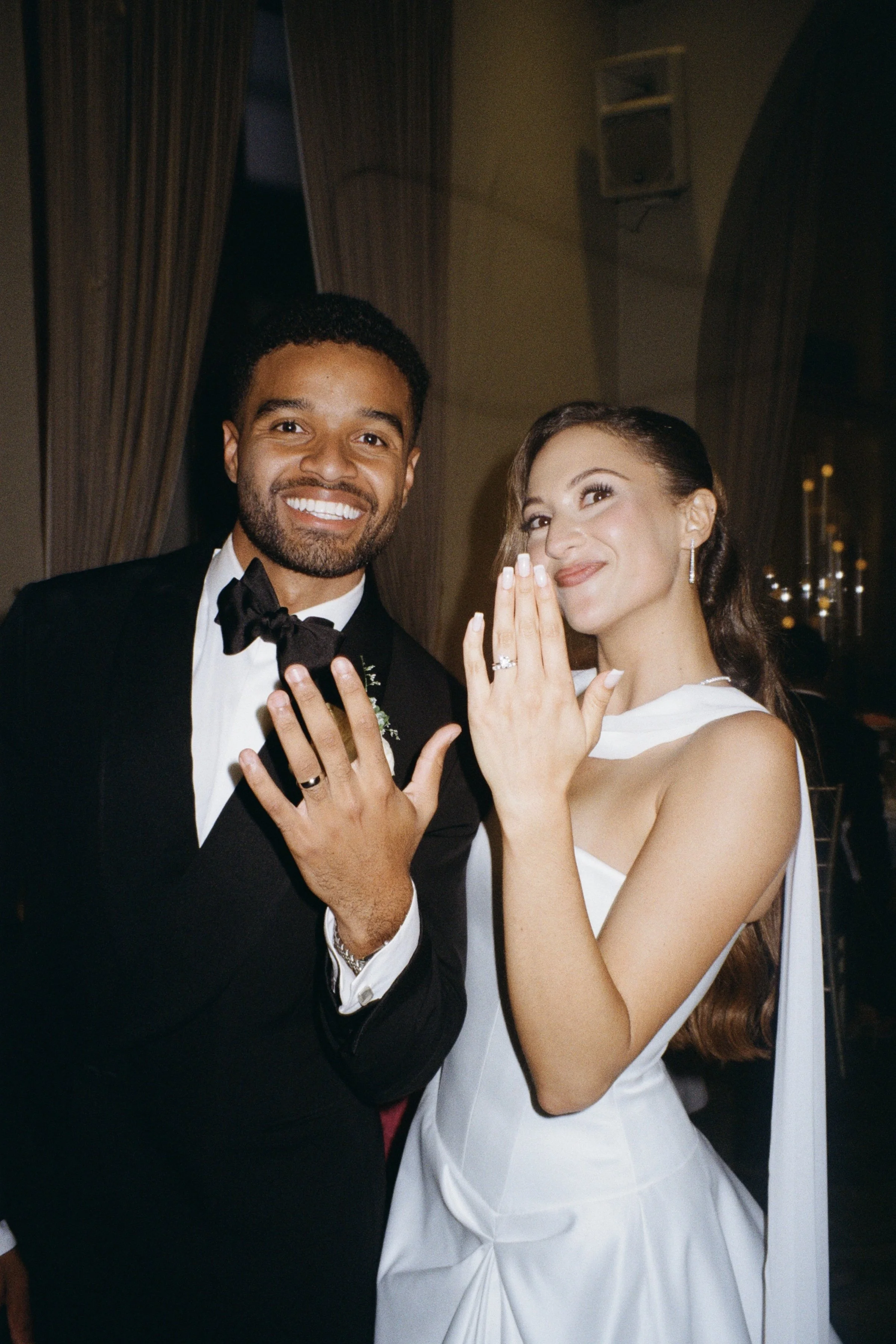 A smiling groom in a black tuxedo and a smiling bride in a white wedding dress showing their wedding rings, with the bride holding her hand near her face.
