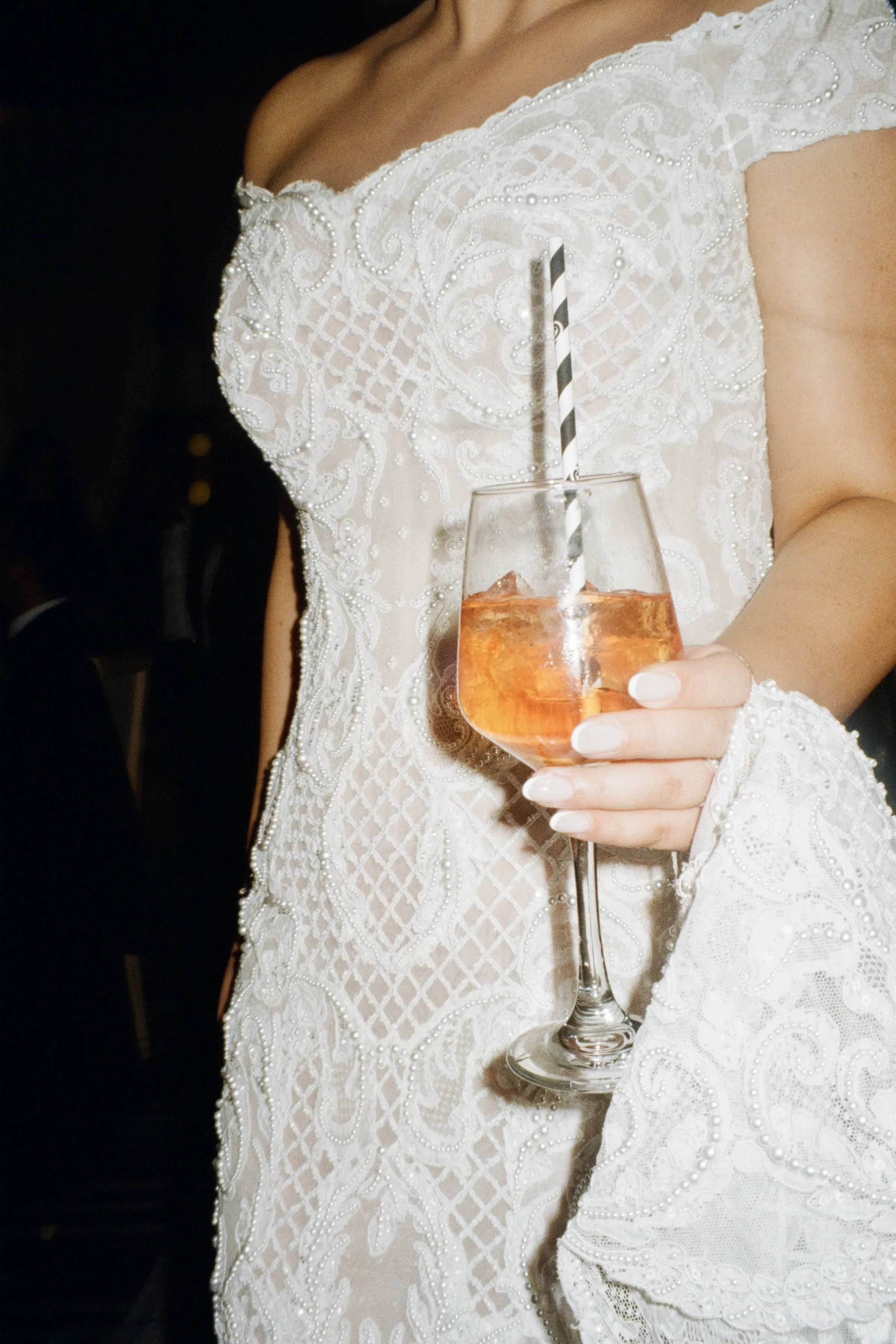 Close-up of a woman in a white lace dress holding a glass of orange-colored drink with ice and a black and white striped straw.