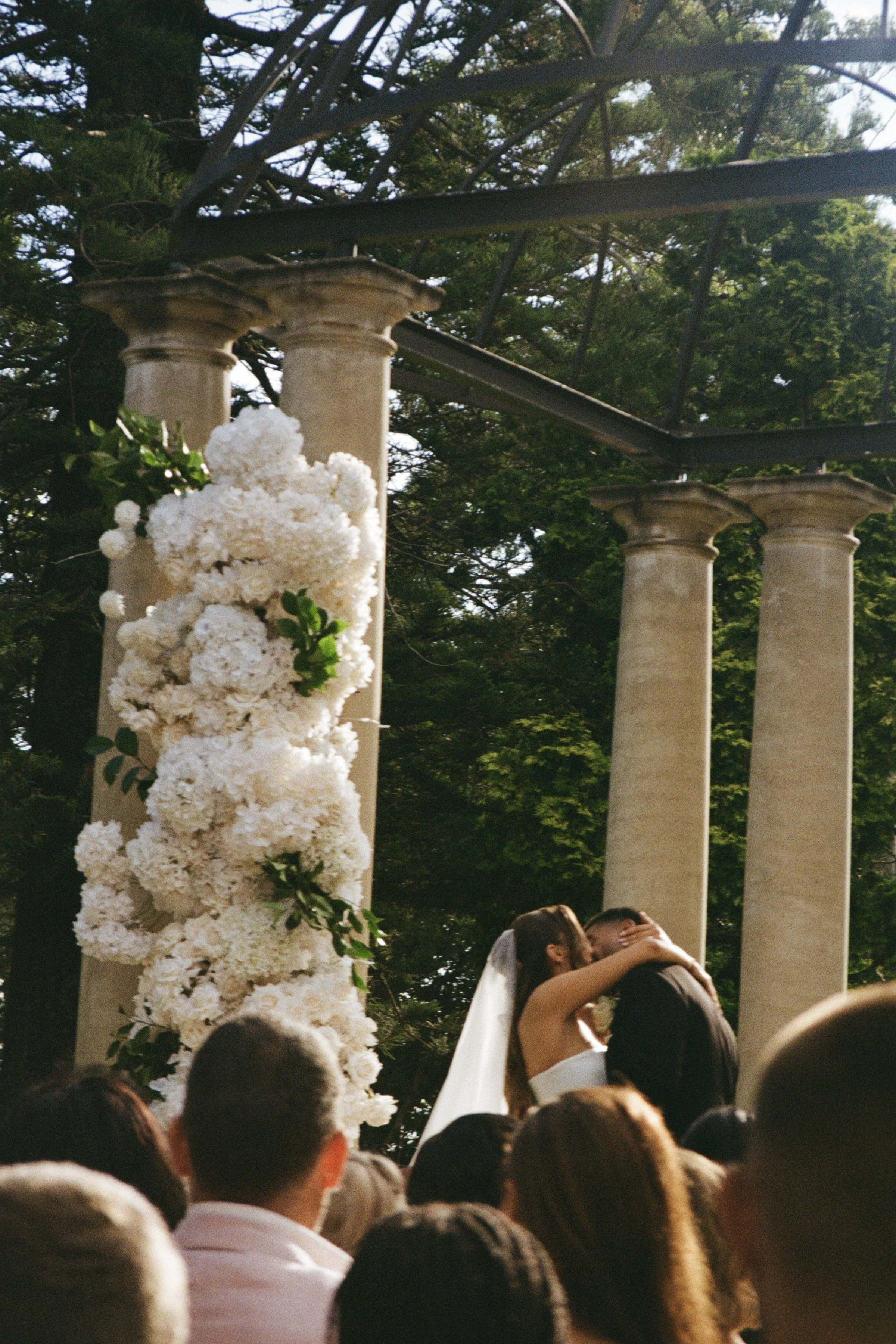 A bride and groom kiss during their wedding ceremony outdoors, surrounded by guests. The setting includes tall pillars and a floral arrangement.