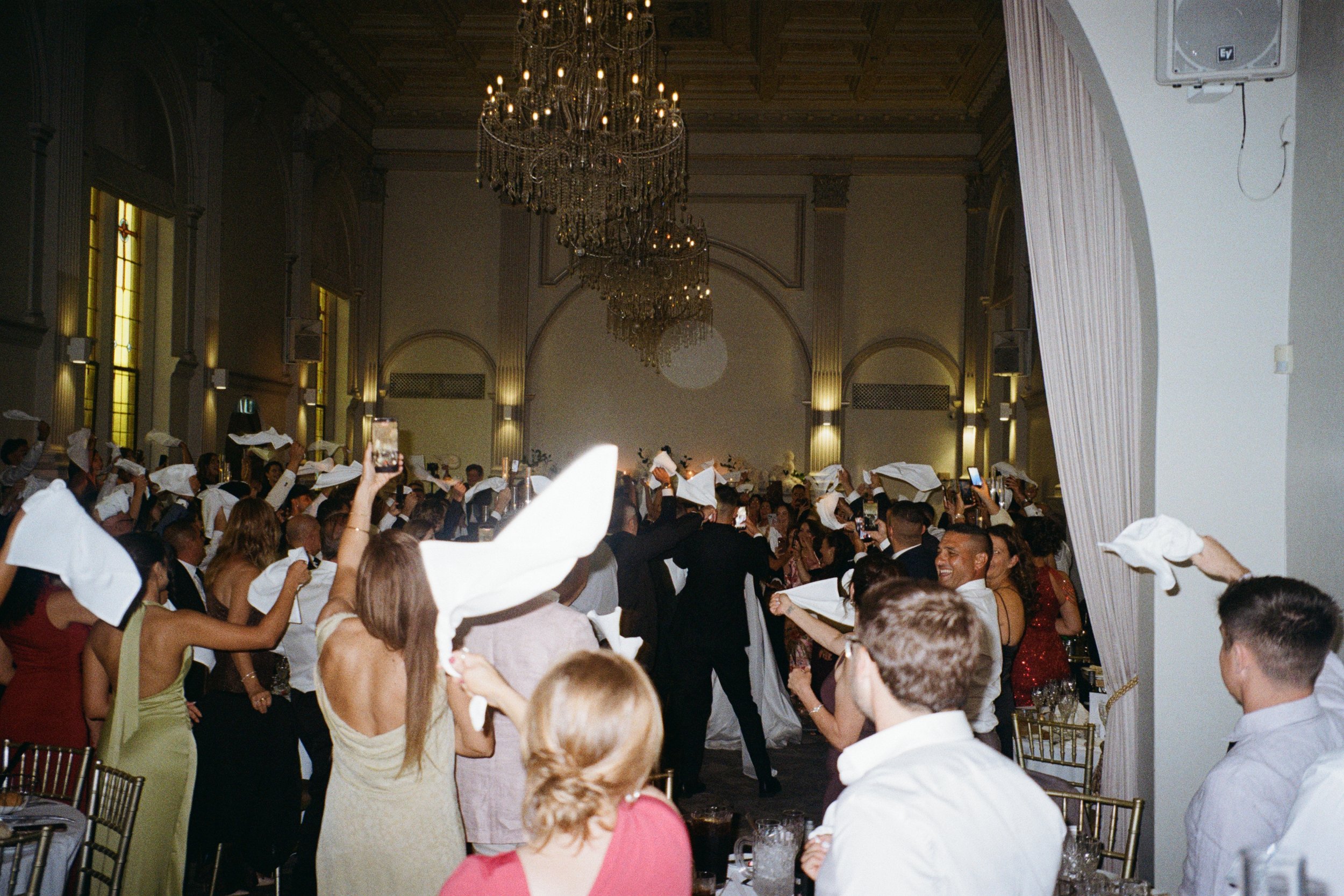 People celebrating at a formal event or party, with some holding napkins in the air, in a large elegant hall decorated with chandeliers and tall windows.