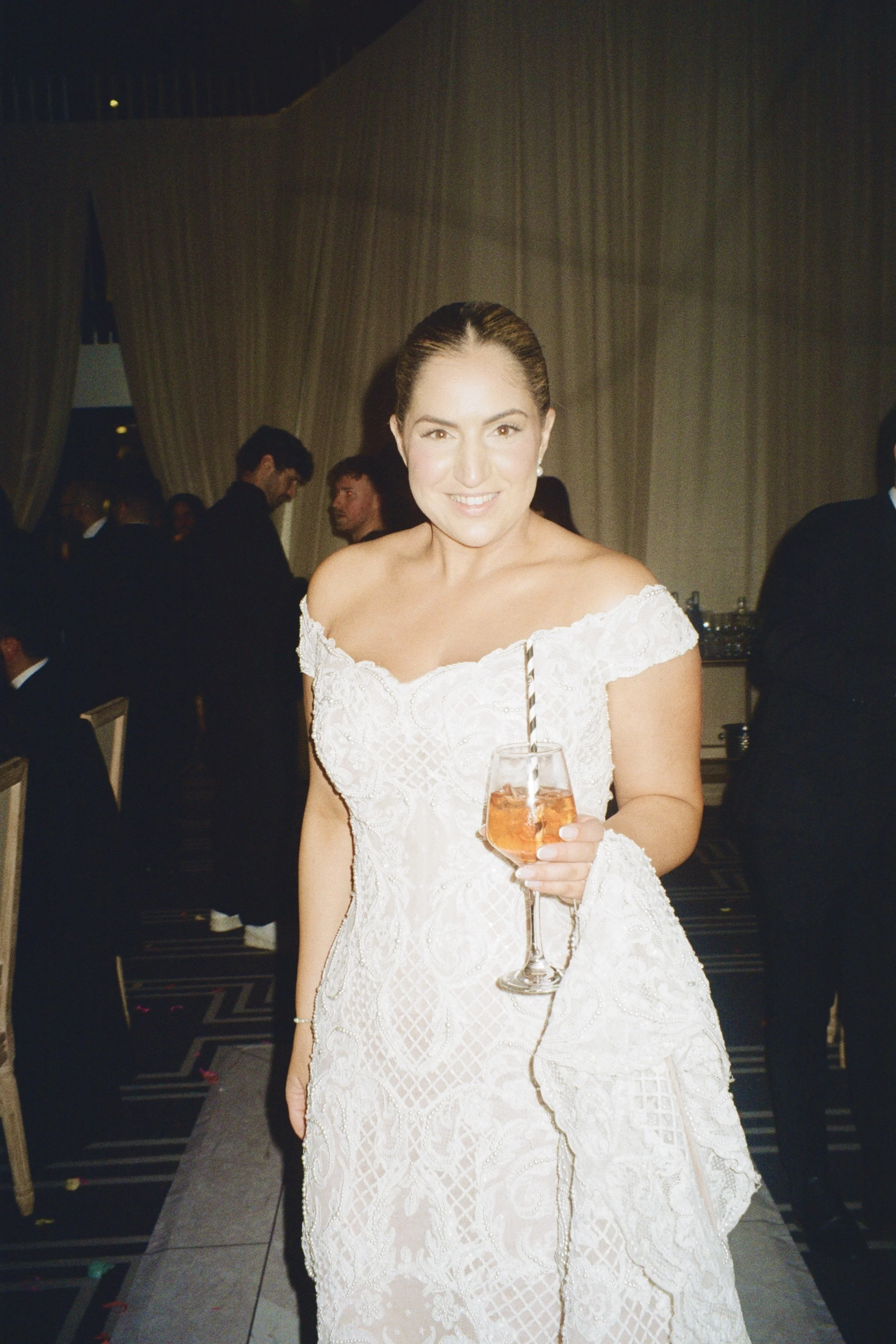 A woman in a white lace wedding dress holding a glass of orange-colored drink with a striped straw, smiling at the camera at an indoor event with people in formal attire and a curtain in the background.