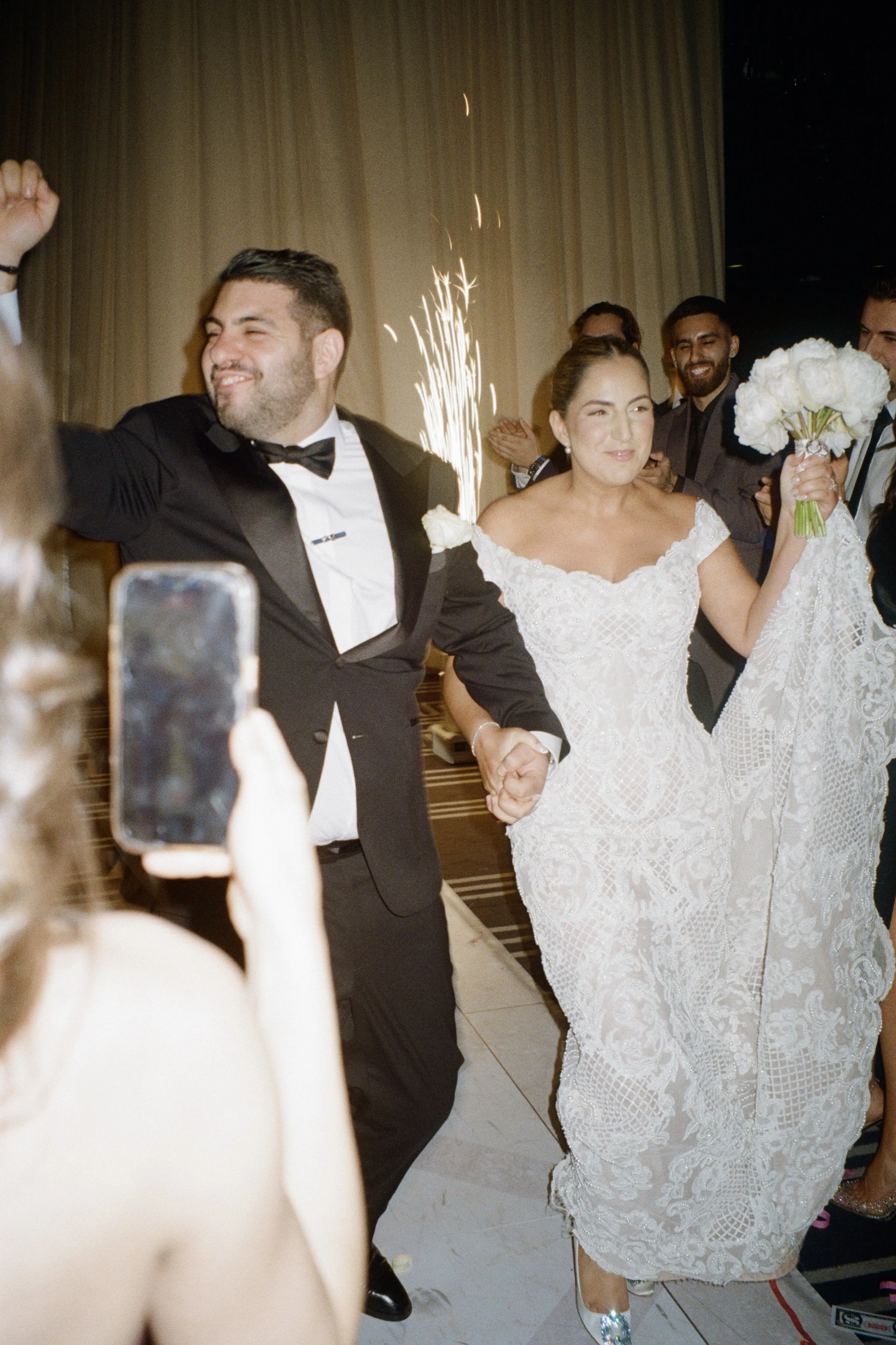 A wedding celebration with a bride in a white lace dress holding a bouquet, a groom in a tuxedo, and guests clapping and taking photos, with a sparkler in the background.