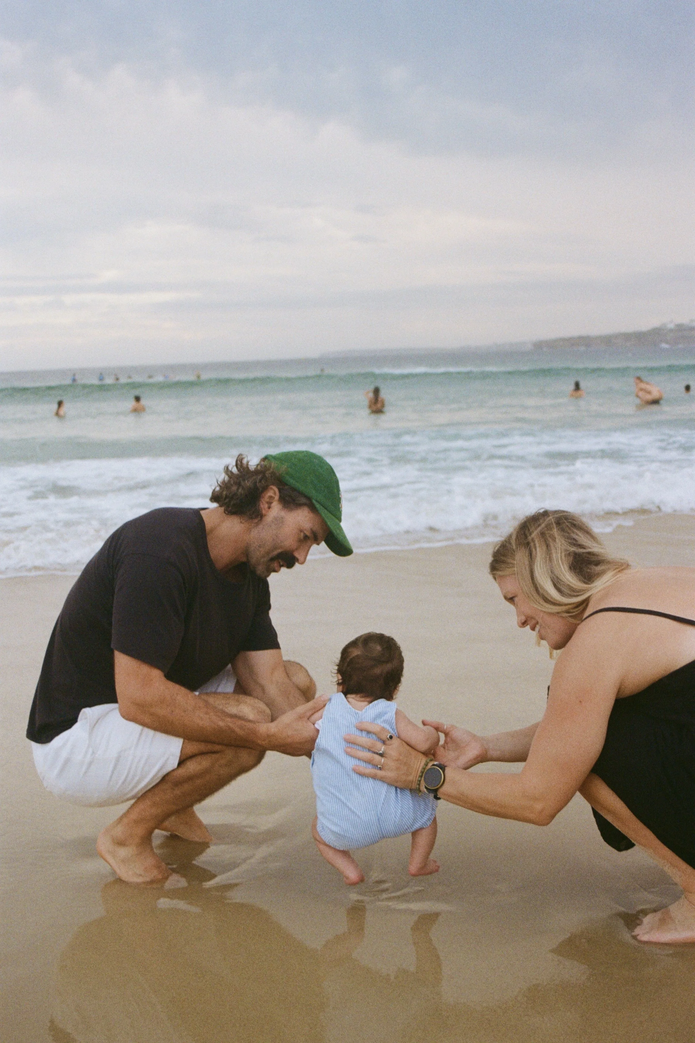 A family on the beach, with a man, woman, and baby, all smiling and happily supporting the baby as it stands on the sand near the water.