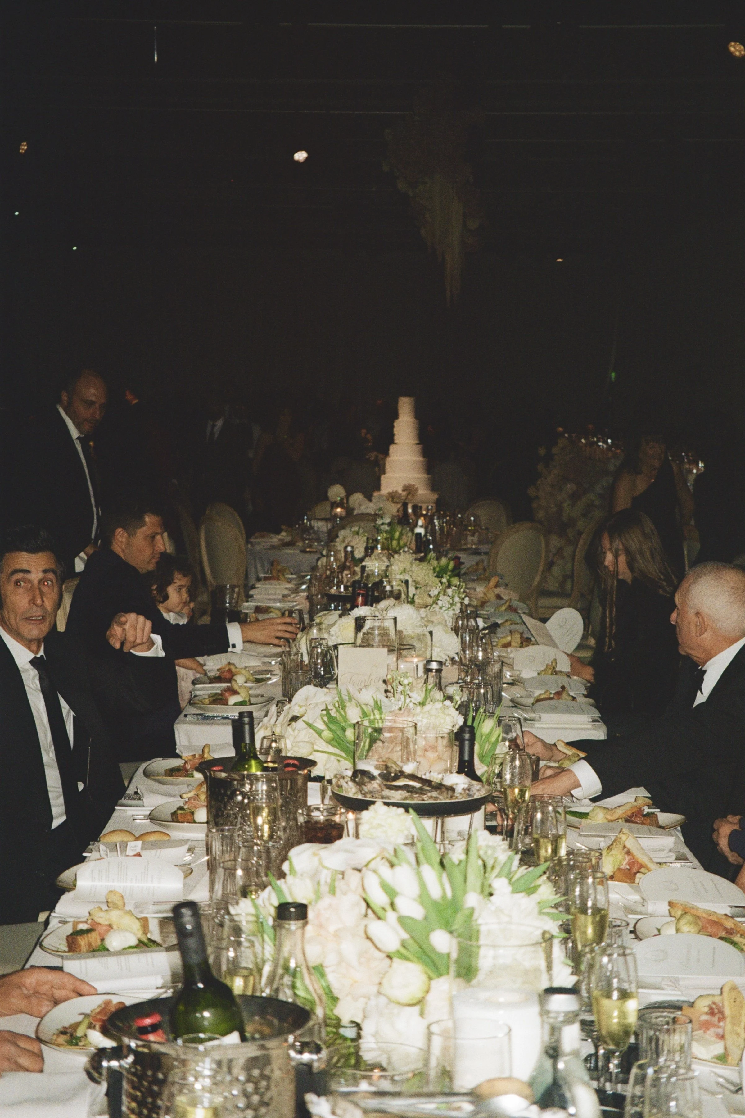 A formal dinner event with guests seated at a long table decorated with flowers, wine bottles, and elegant tableware, with a multi-tiered cake in the background.