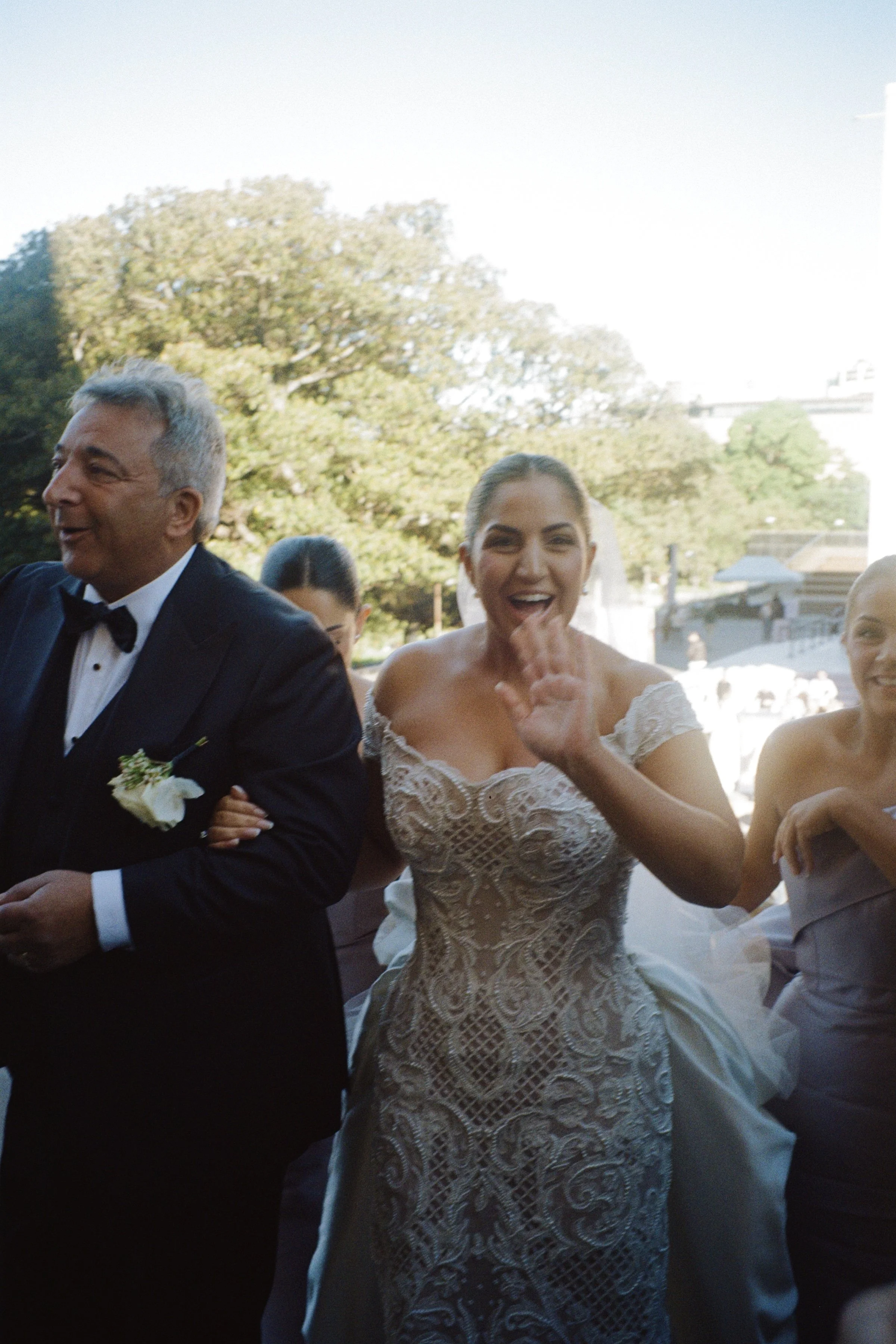 Group of wedding guests outdoors, including a woman in a lace wedding dress and an older man in a tuxedo, smiling and celebrating.