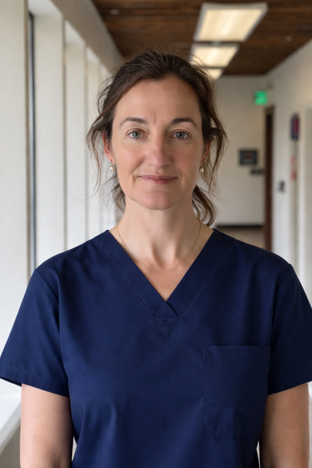Portrait of a woman with brown wavy hair, wearing navy scrubs, standing in a hospital corridor with white walls and wooden ceiling panels.