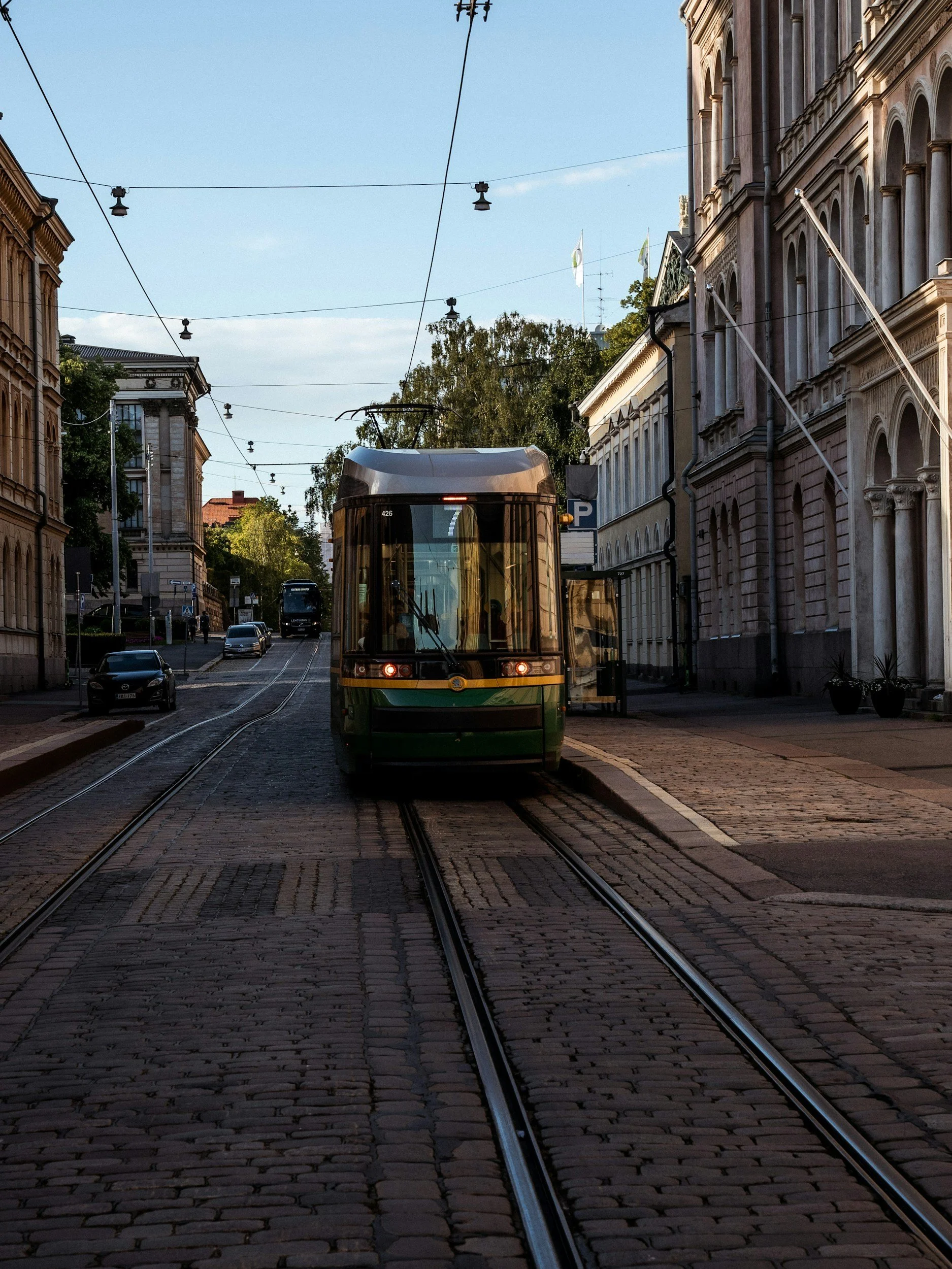 A green tram traveling on cobblestone tracks through a city street with historic buildings on either side and overhead wires for the tram.