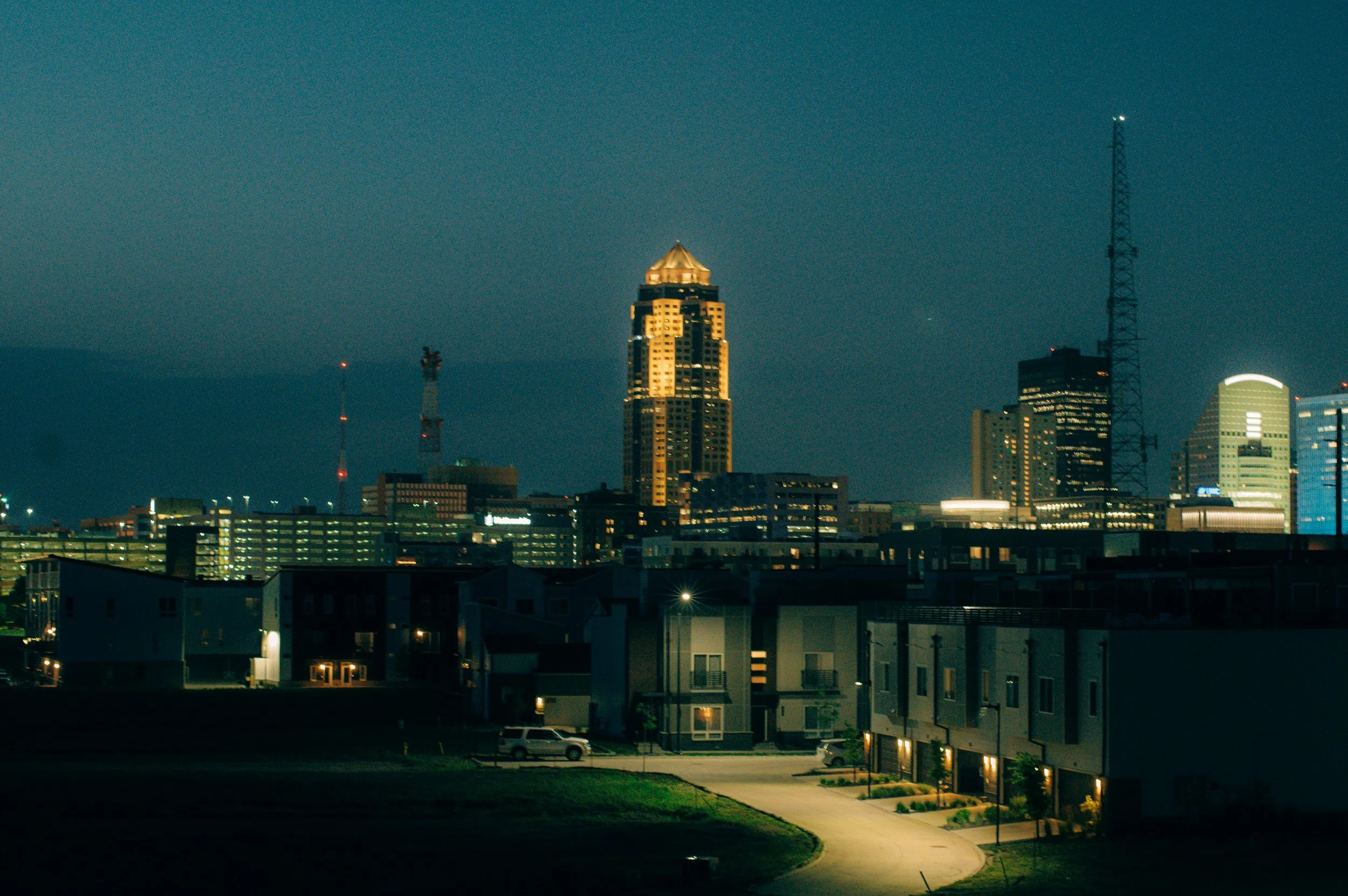 Nighttime cityscape with illuminated buildings and skyscrapers, some residential houses in the foreground, and a clear dark sky.