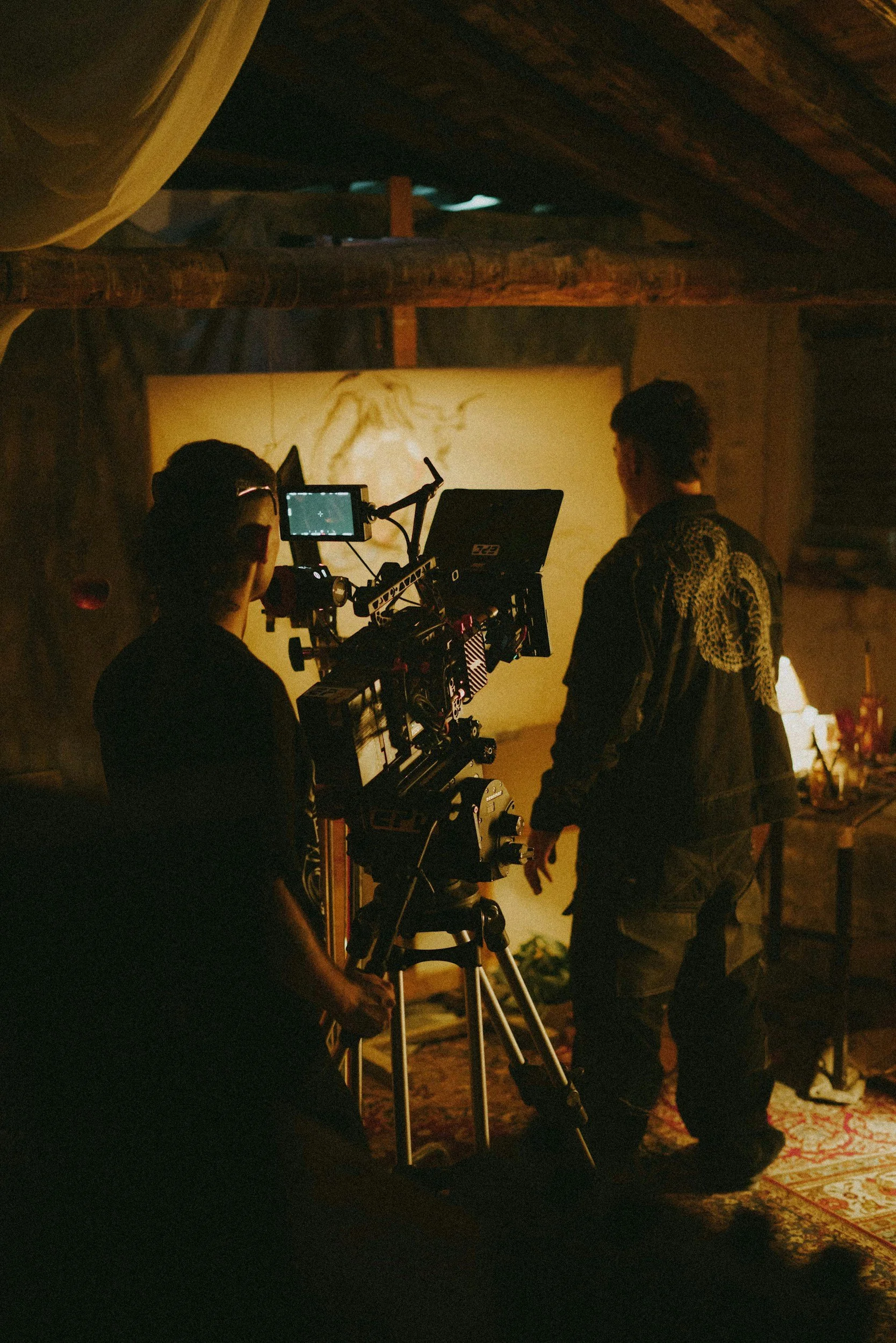Two individuals operating a professional film camera on a tripod in a dimly lit, rustic interior with wooden beams and a canvas hanging on the wall in the background.