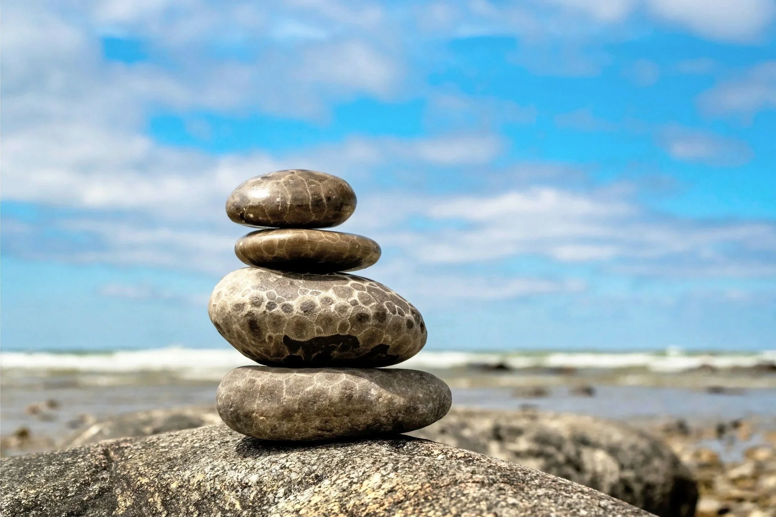 Stack of four smooth stones arranged from largest at the bottom to smallest at the top on a rocky beach with the ocean and blue sky with clouds in the background.