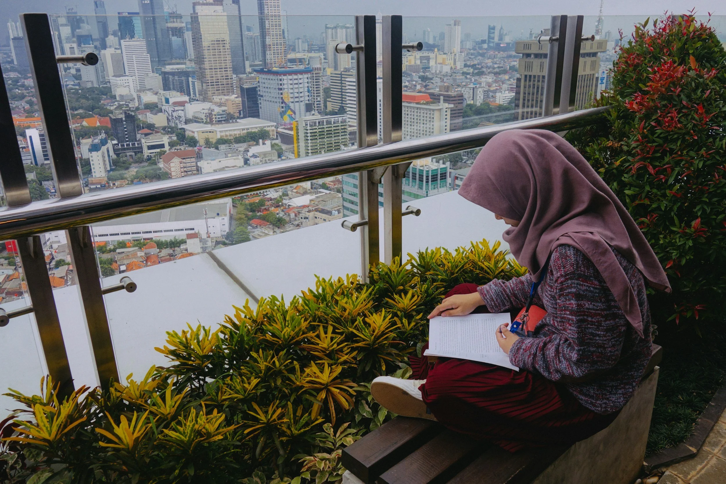 A woman wearing a hijab is sitting on a bench, reading a book, with a city skyline in the background.