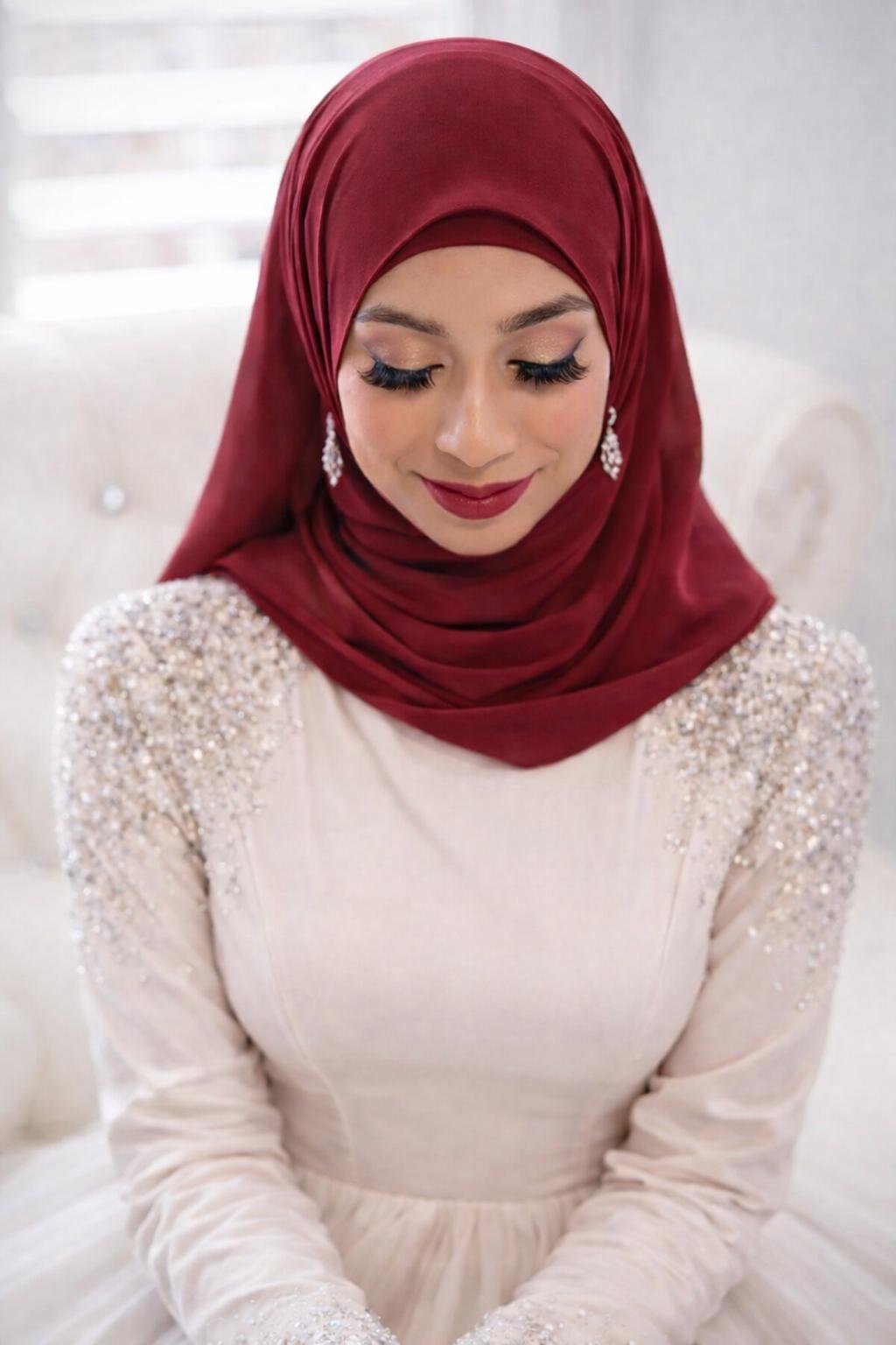 A woman wearing a red hijab and an elegant, cream-colored dress with beaded shoulders, smiling with eyes closed.