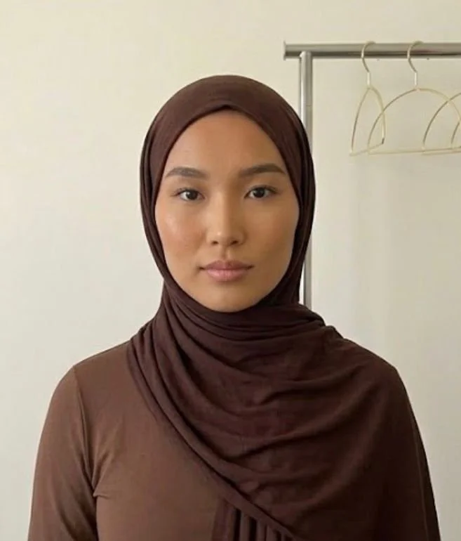 A woman wearing a brown hijab and matching top standing in front of a plain wall with a metal rack and empty hangers in the background.
