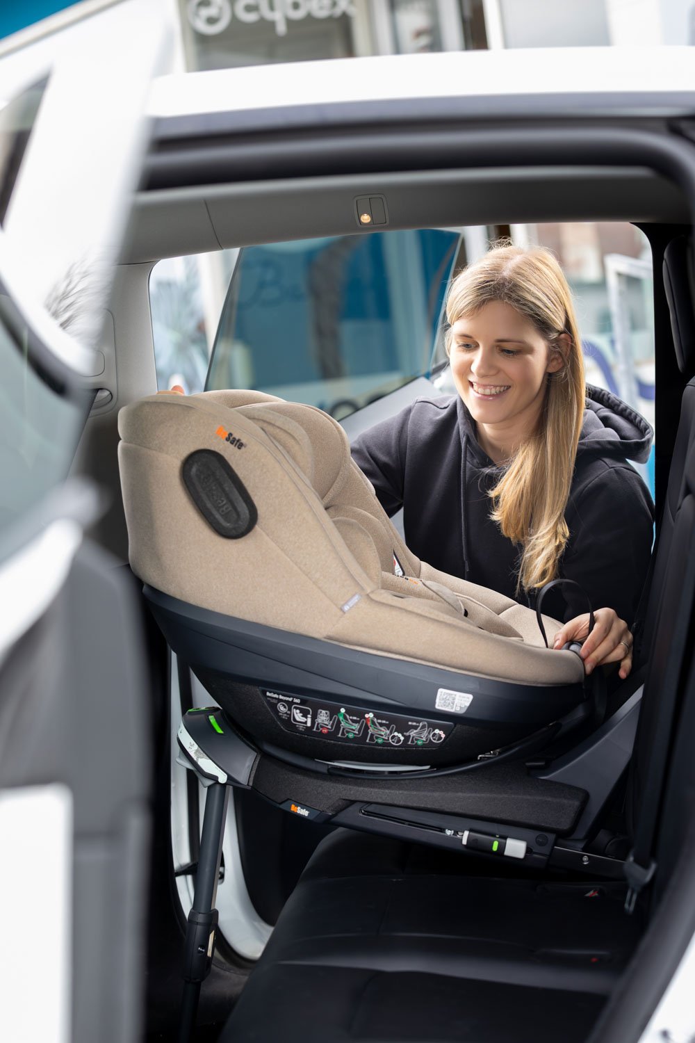 Eine junge Frau installiert einen in beige gehaltenem Autositz im Fond eines Autos.