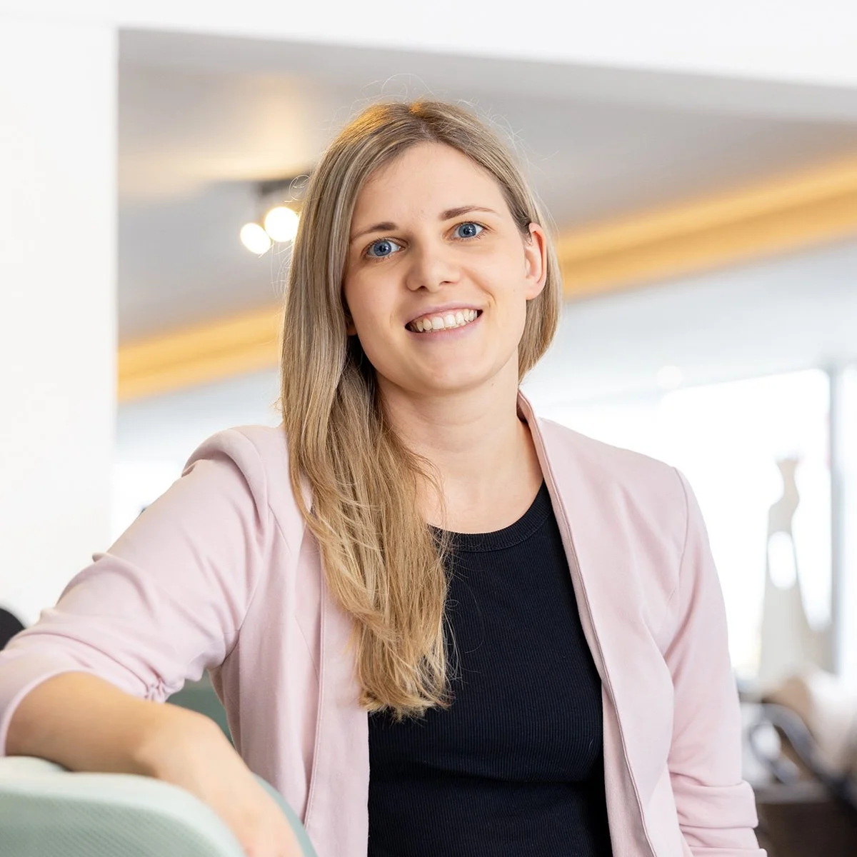 Junge Frau mit blonden Haaren und blauen Augen lächelt in einem hellen Raum.