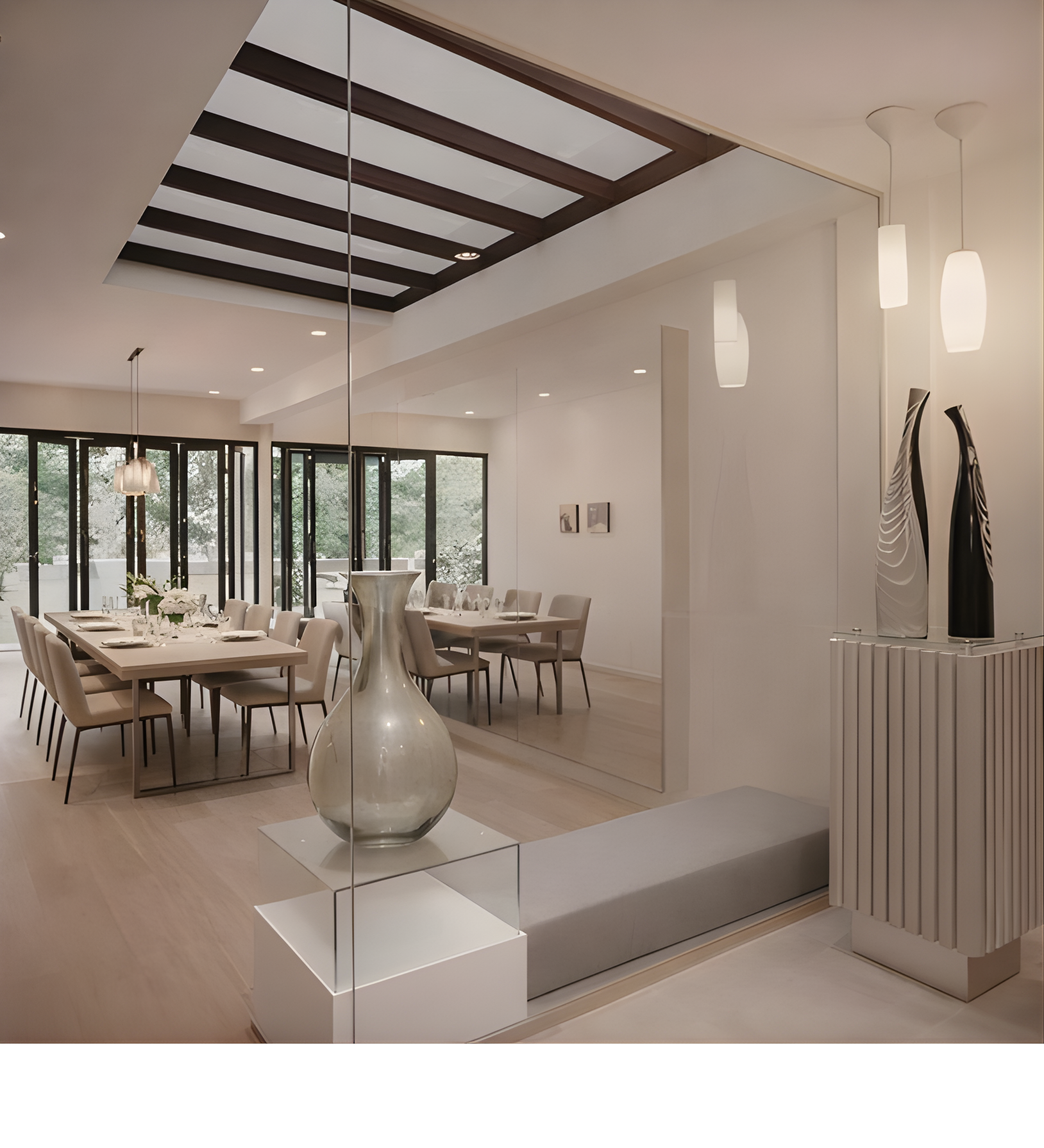 Modern dining room with a large white table, beige chairs, and a white decorative vase on a white pedestal, separated by a mirrored wall, with pendant lights and large windows providing natural light.