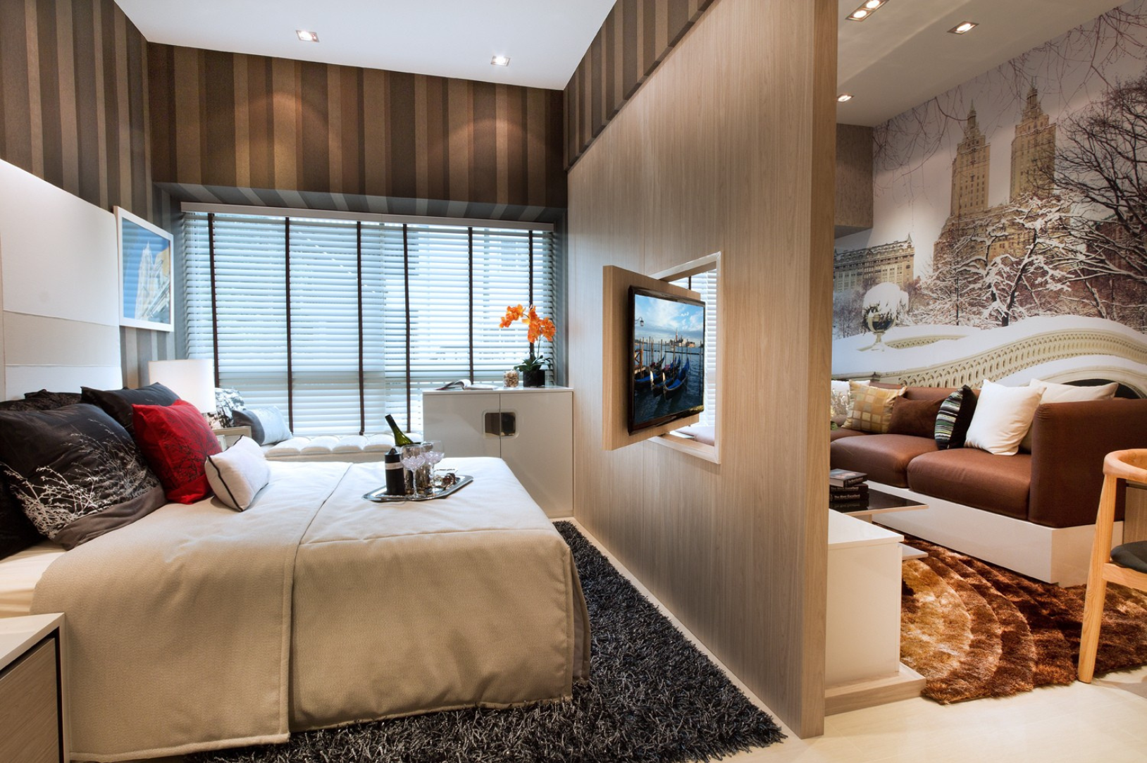 Modern apartment bedroom with beige bed, black and red pillows, champagne bottle with glasses on tray, window with blinds, adjacent living area with brown sofa and snow-themed mural.