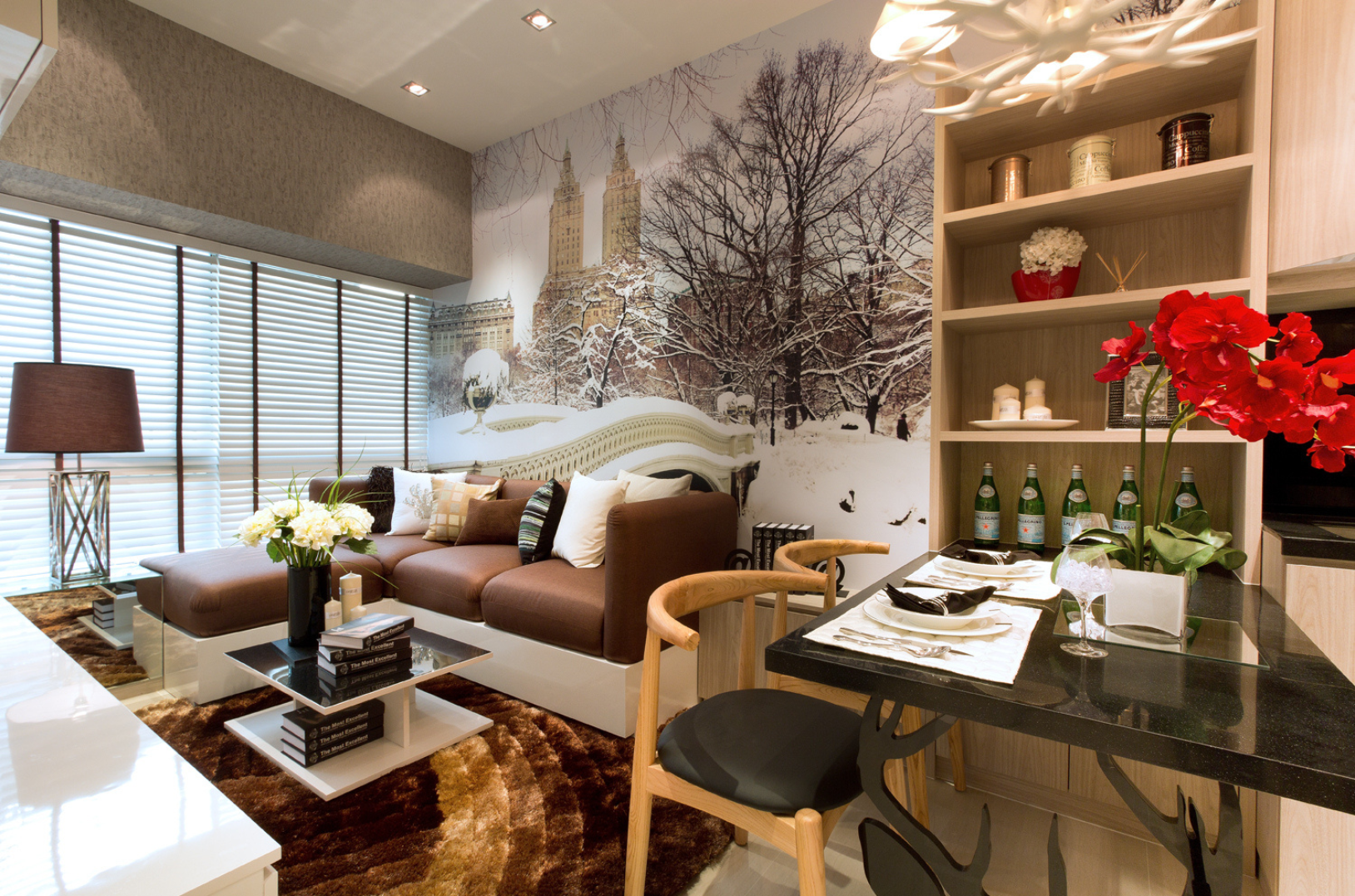 A living room with a brown sectional sofa, a coffee table with books and flowers, a large window with blinds, a wall mural of a snowy park, and a dining area with a black table set for two, red flowers, and shelves with decor.