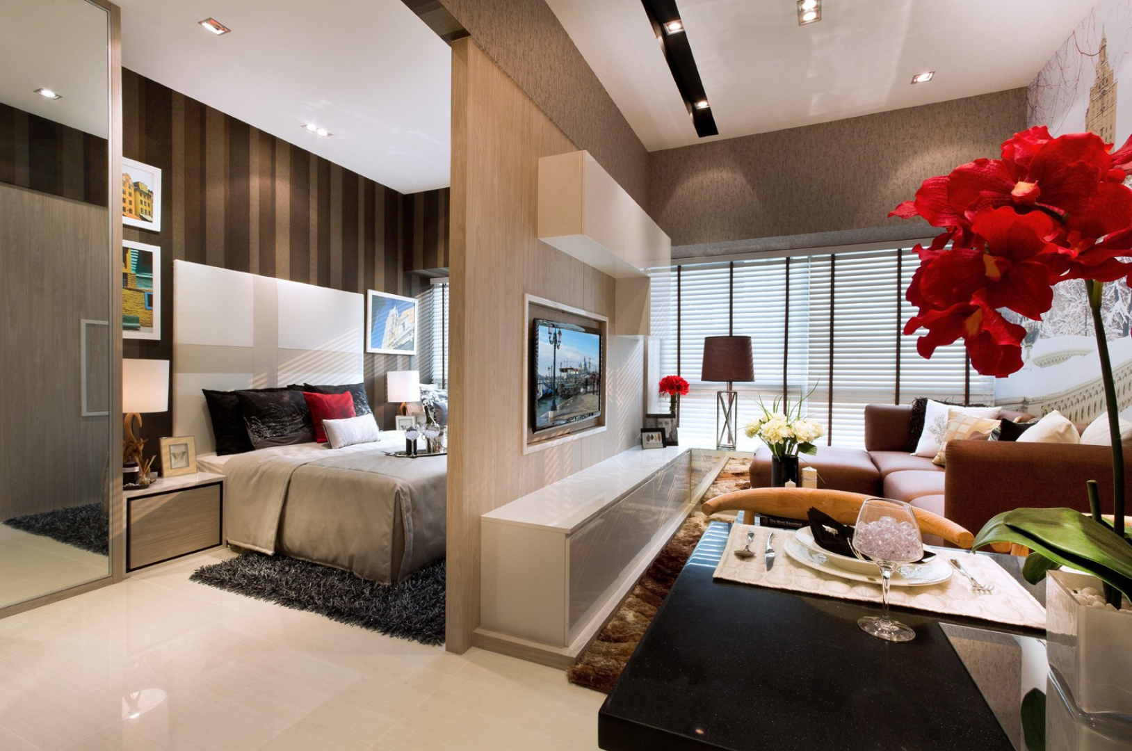 Modern living room and bedroom separated by a partial wall, featuring a bed with black and red pillows, a wall-mounted TV, a brown sofa with decorative pillows, and a dining area with red flowers, a wine glass, and a table setting.