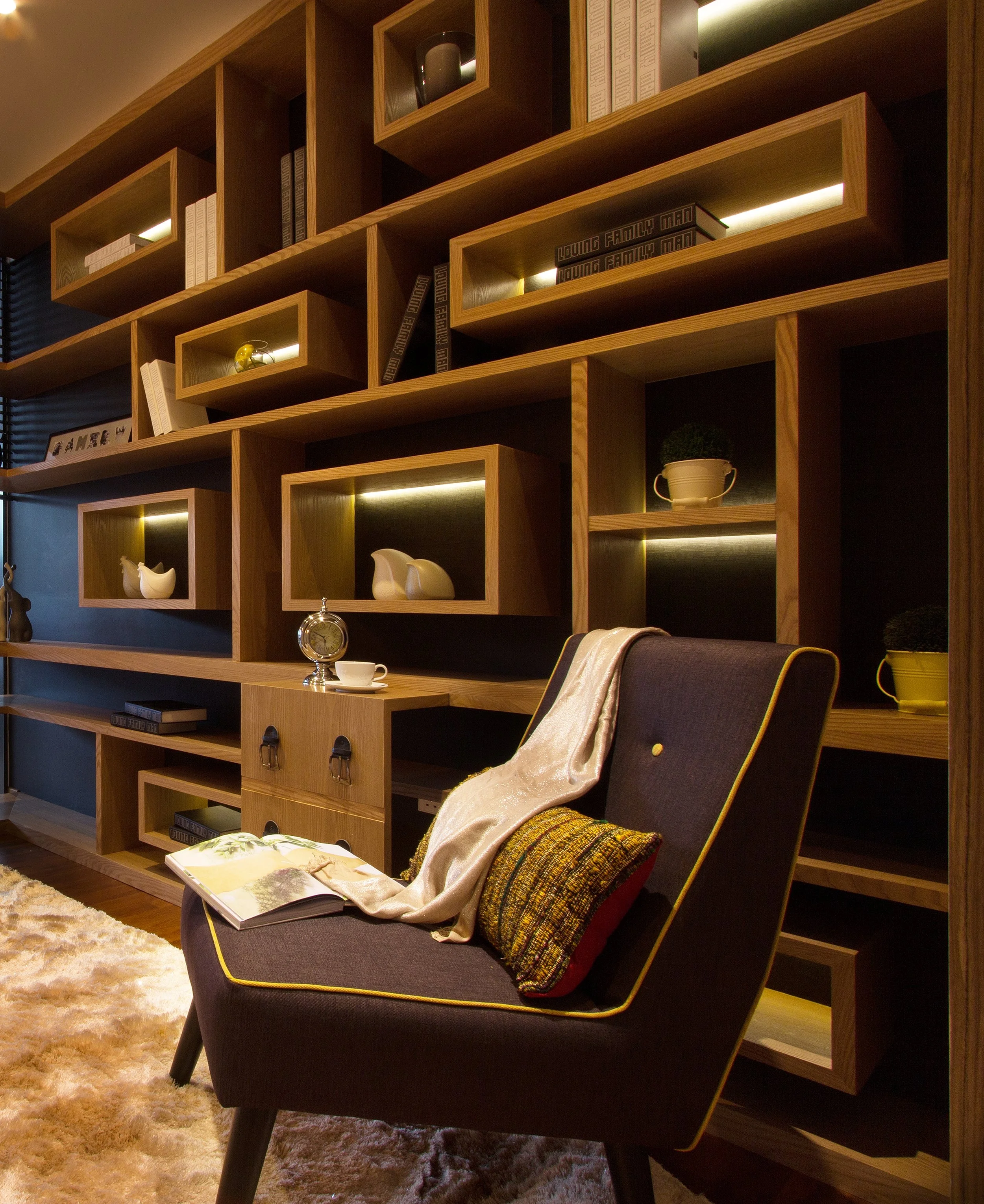 A modern living room with a large wooden bookshelf, a black armchair with yellow piping, and decorative items including books, vases, and a clock. A cozy blanket is draped over the armchair, and a book is open on the armrest. The floor is covered with a shaggy, cream-colored rug.