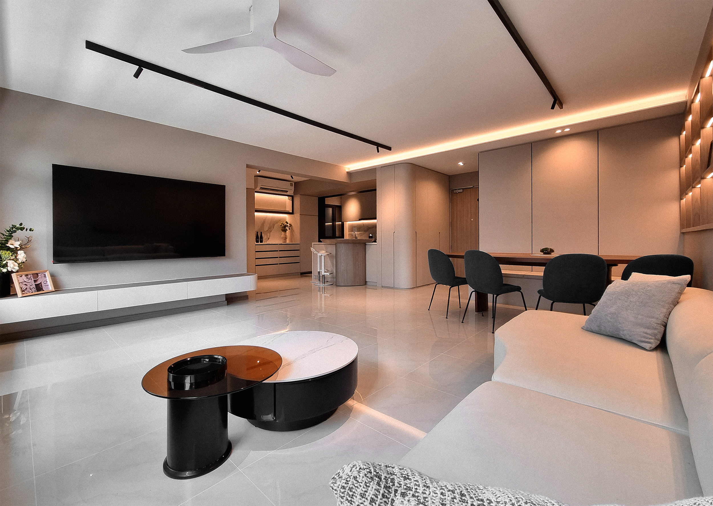 Modern living room with large flat-screen TV, beige sofa, black and orange coffee tables, dining area with black chairs, and minimalistic decor and lighting.