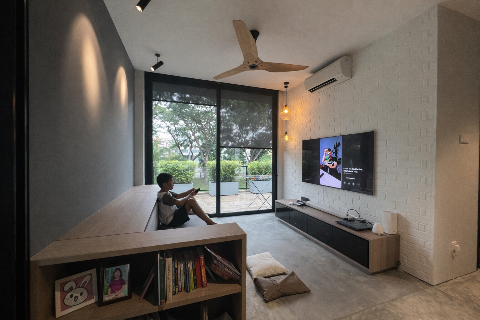 A young boy sitting on a built-in wooden bench in a modern living room, looking at a smartphone. The room features a white brick wall with a wall-mounted TV displaying a colorful image, a black entertainment unit, a large glass sliding door opening to a balcony with greenery outside, and is decorated with hanging pendant lights and a ceiling fan.