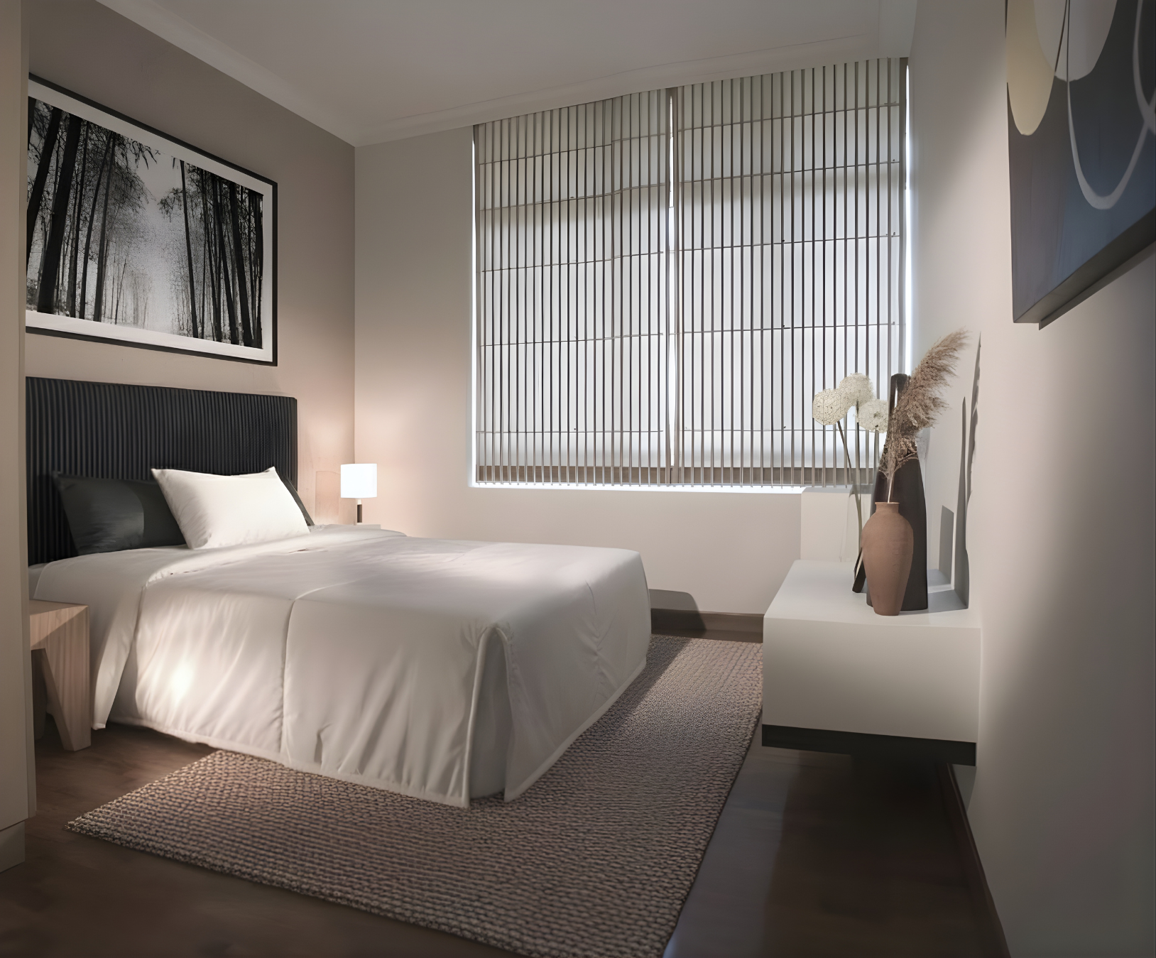 A modern bedroom with a neatly made bed, black headboard, white bedding, and pillows. A small bedside table with a lamp is beside the bed. The room has a large window with wooden blinds, and a white side table with decorative vases and dried plants. A black and white forest artwork hangs on the wall above the bed.