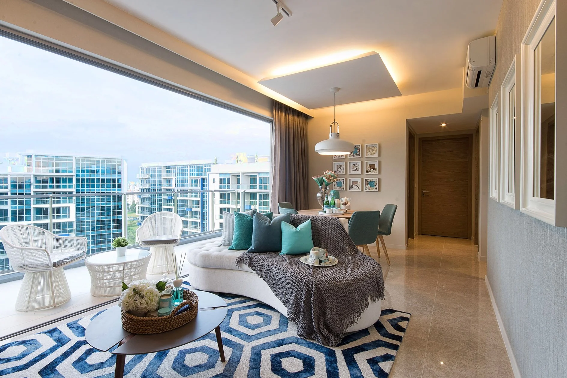Living room with a large window overlooking tall modern apartment buildings, decorated with blue and white cushions and a geometric rug, with a small round table, wicker chairs, and interior decor in a contemporary style.