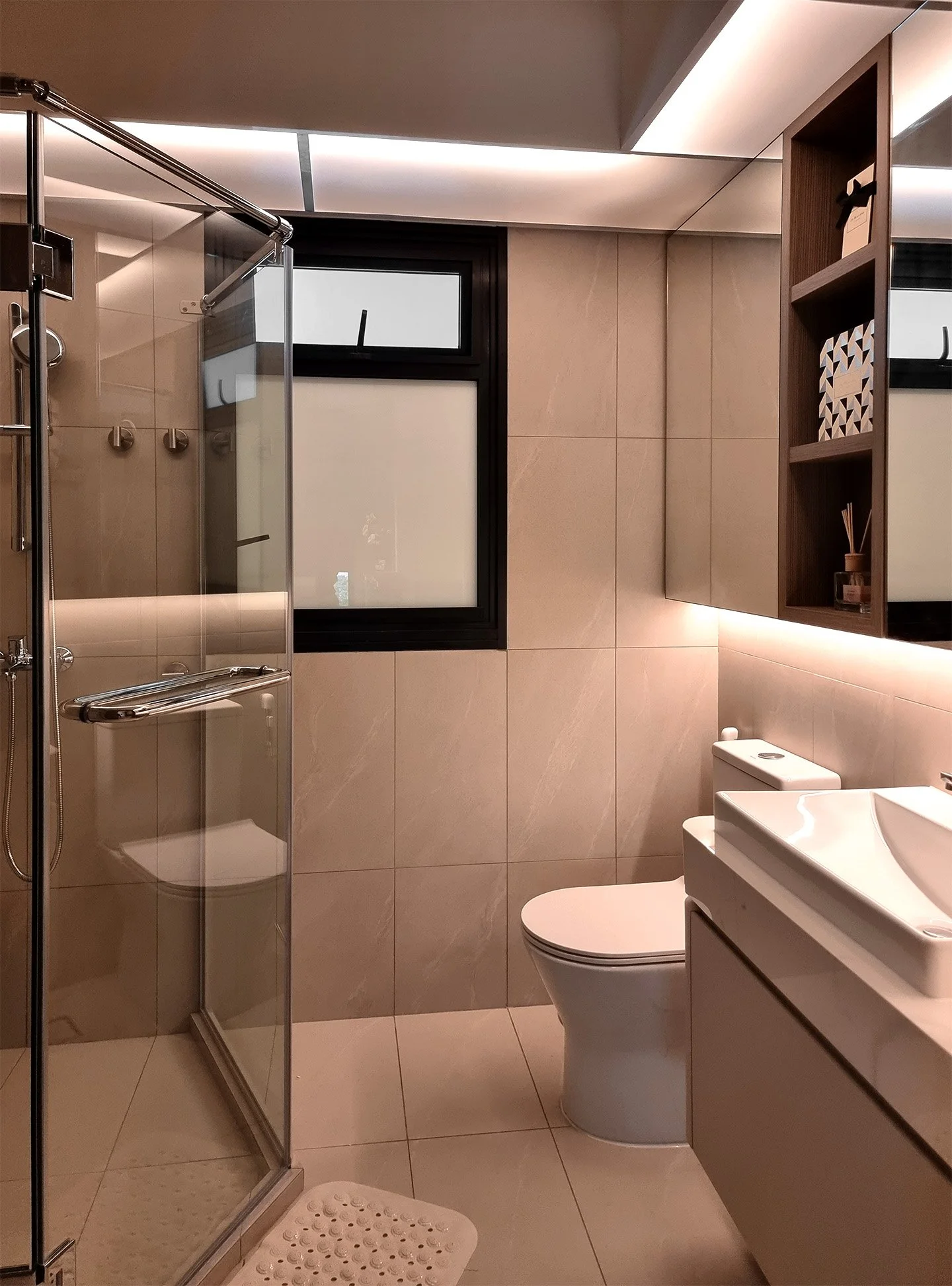 Modern bathroom with beige tiled walls, a frosted window, a glass shower stall, a white toilet, and a sleek white sink with a wooden cabinet. There is a wall-mount shelf with decorative items and a bath mat on the floor.