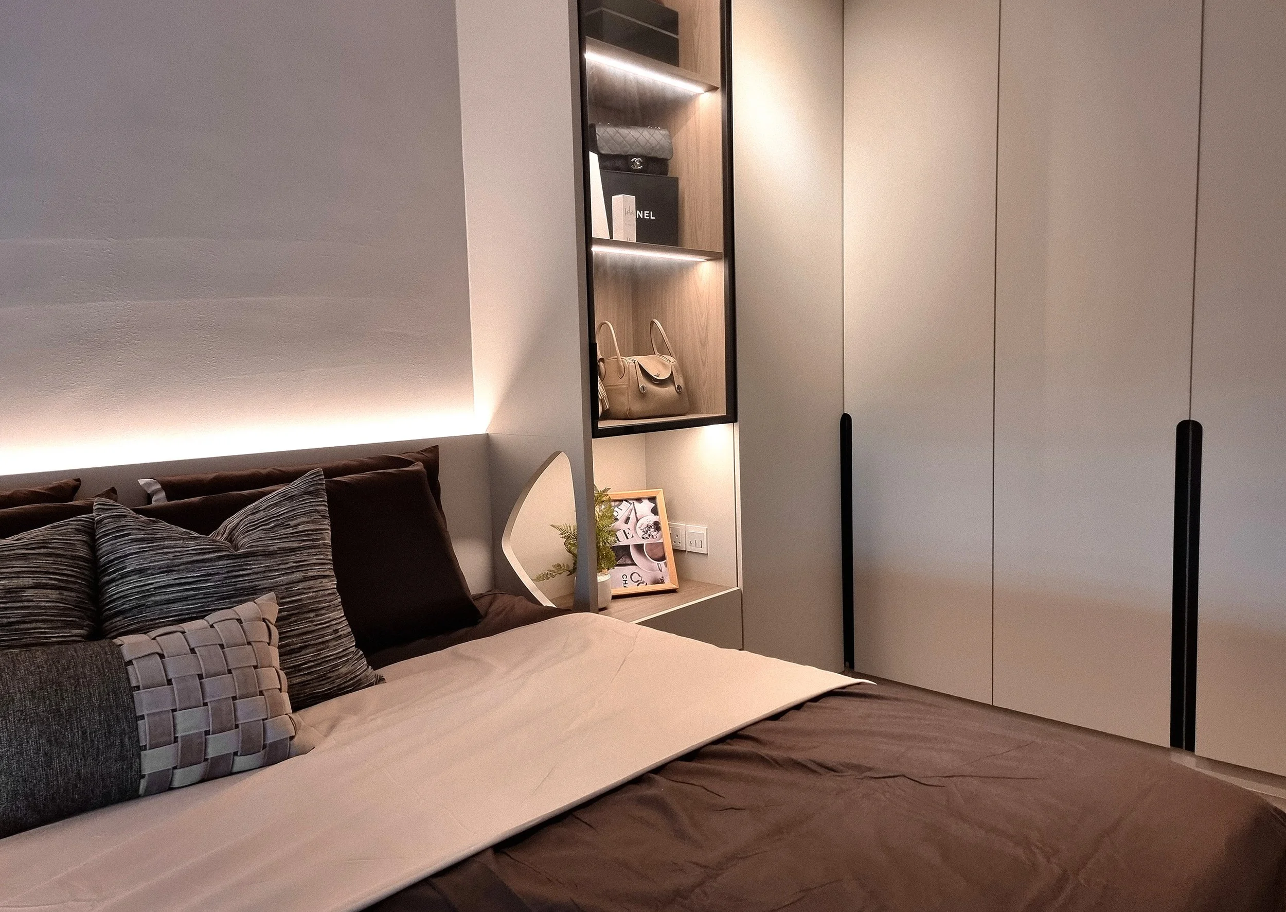 A neatly made bedroom with a bed, multiple pillows, and a white bedside table with a framed picture and a small plant. To the right, there is a tall built-in closet with closed doors and black handles. Next to the closet, there is an open shelving un