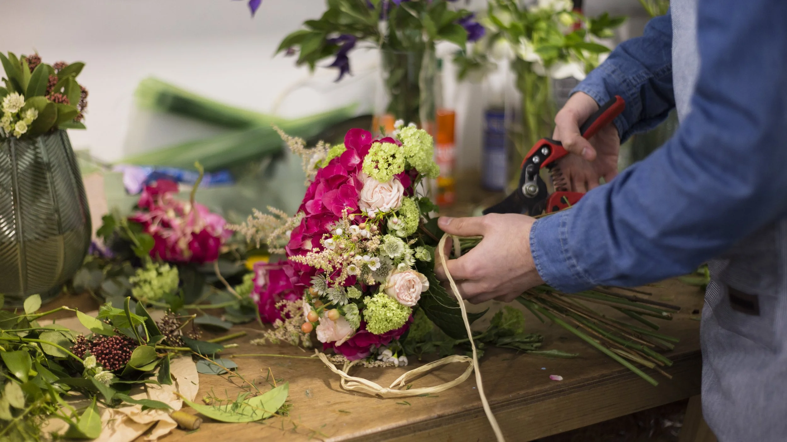 Person arranging a floral bouquet with pink, white, and green flowers in a workshop setting.