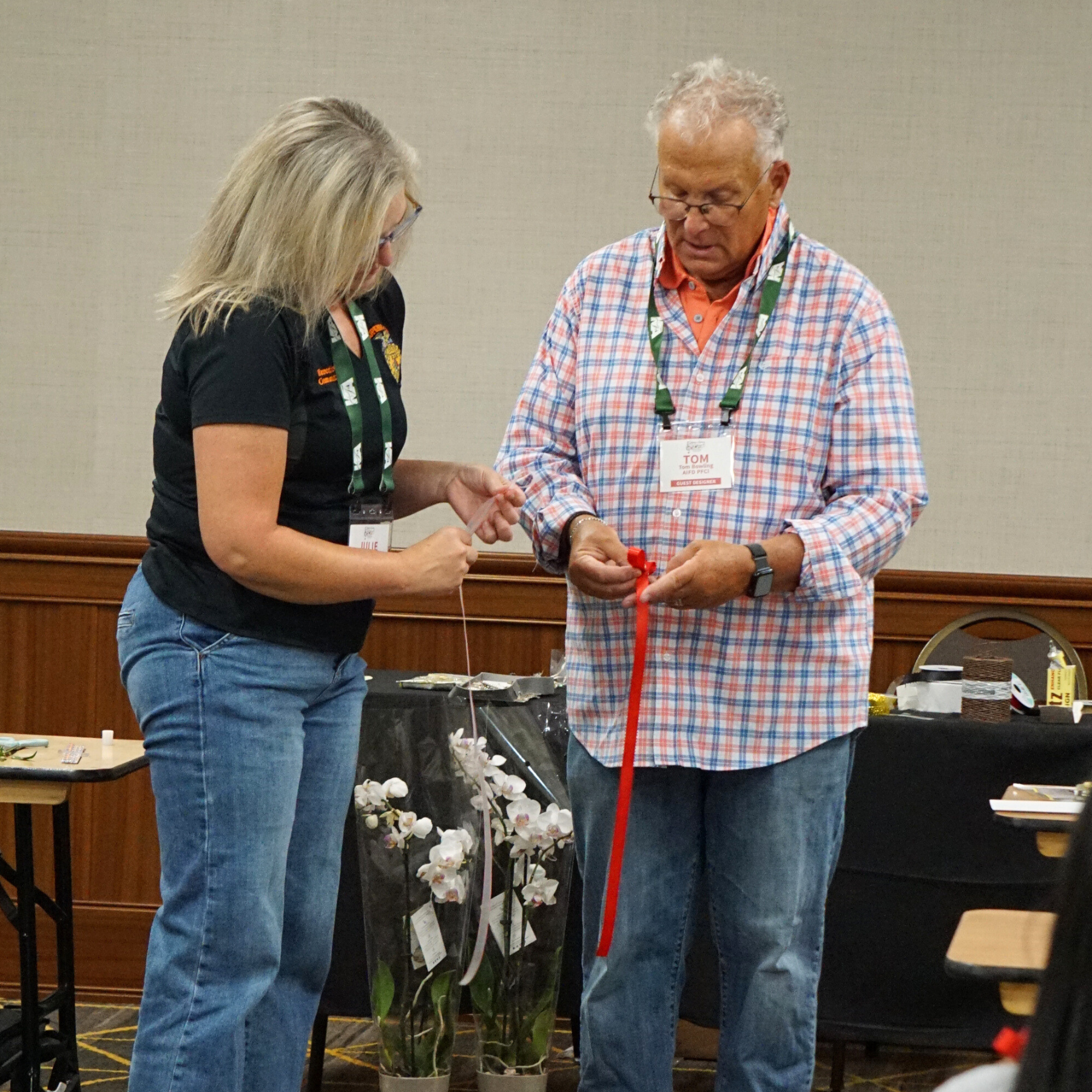Two people, a woman and a man, are standing and looking at something together. The woman has blonde hair, is wearing glasses, a black shirt with a logo, and jeans. The man has curly gray hair, is wearing glasses, a plaid shirt over an orange polo shirt, and jeans. They both have conference badges around their necks. There are orchids and other items on the table behind them, and the setting appears to be indoors with a beige wall in the background.