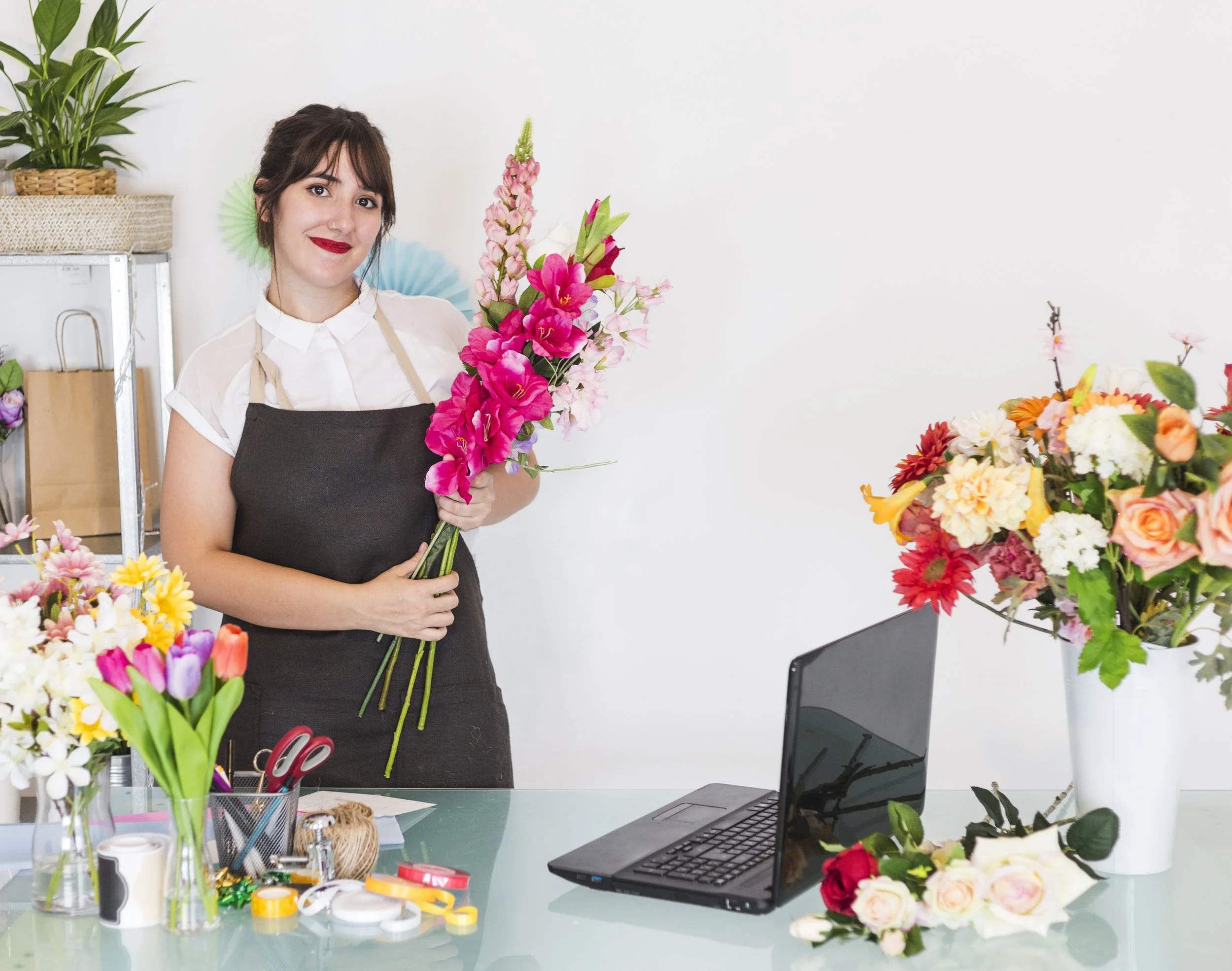 A woman with dark hair and red lipstick holding a large bouquet of pink and white flowers in a room decorated with various colorful flowers and green plants, with a laptop and office supplies on a desk.