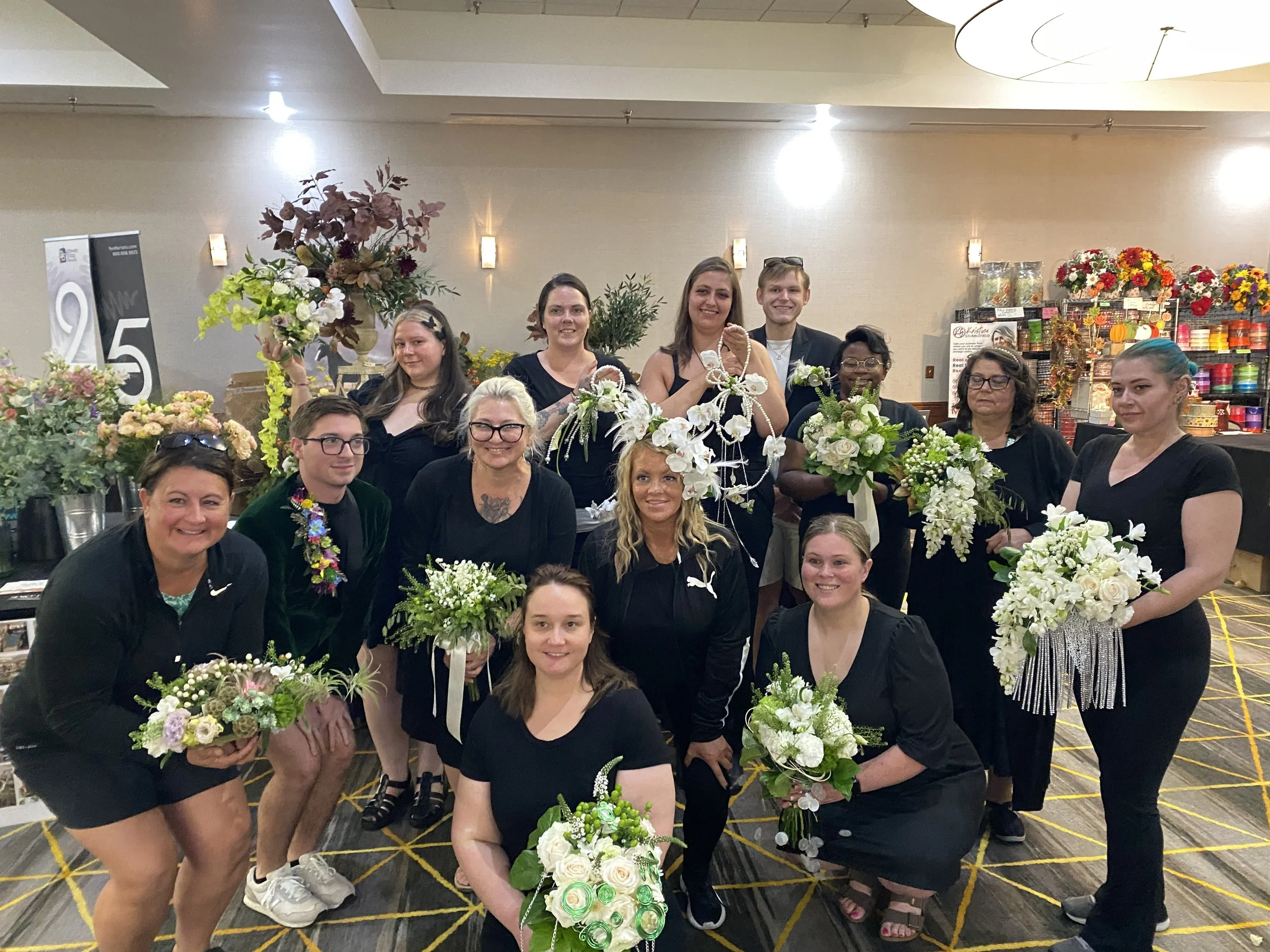 Group of people gathered at a floral event, holding bouquets and surrounded by various flower arrangements, indoor setting with beige walls and ceiling lights.