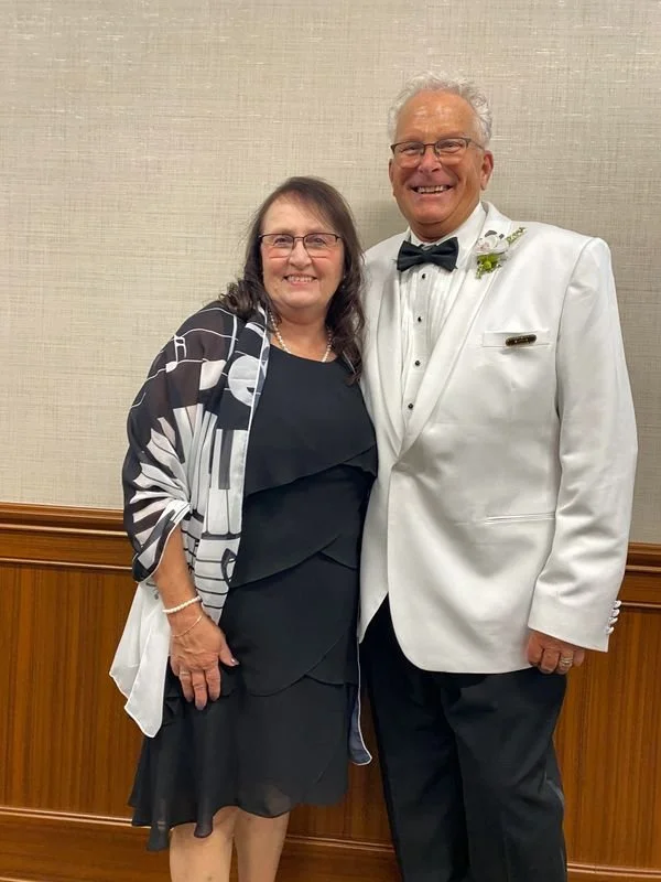 A smiling woman in a black dress and patterned shawl, standing next to a smiling man in a white tuxedo with a black bow tie, against a beige wall with wood paneling.