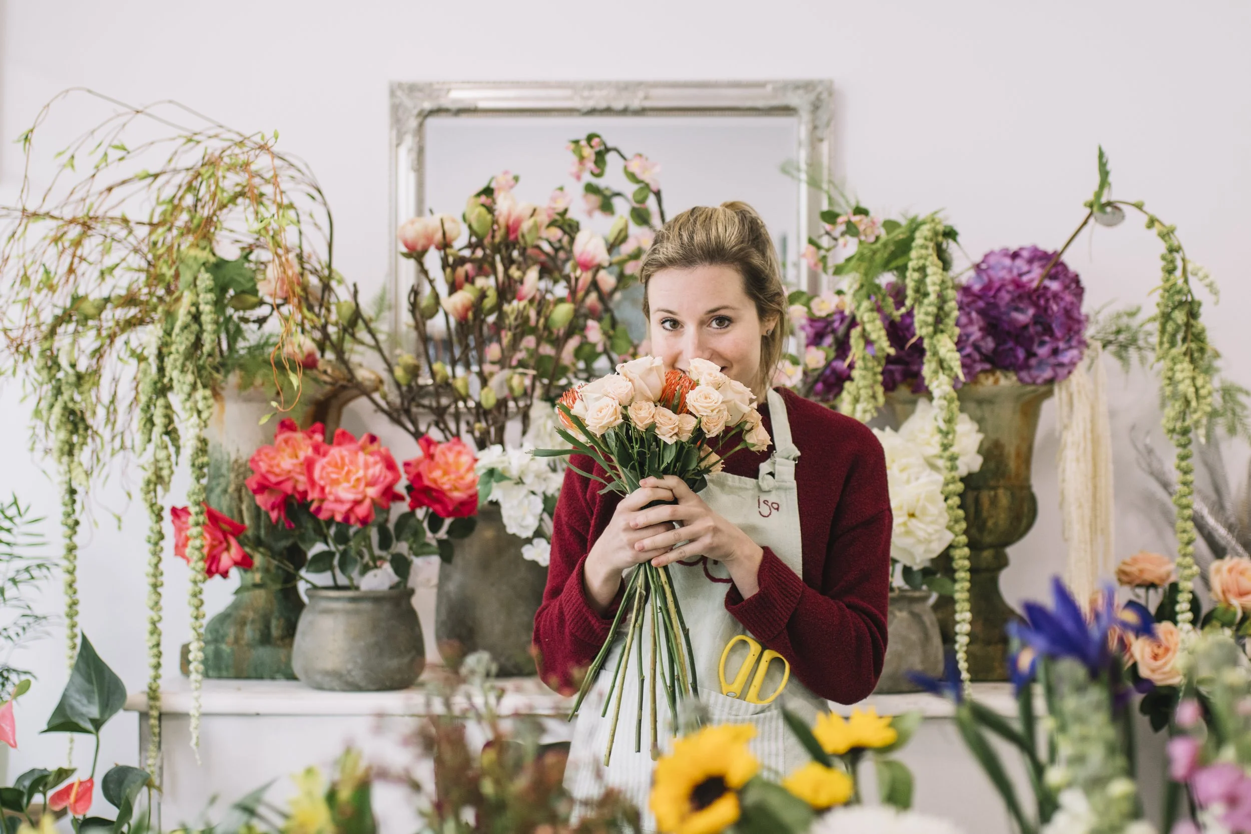 A woman in a florist shop holds a bouquet of pale pink roses and orange flowers, surrounded by various colorful flowers and plants.